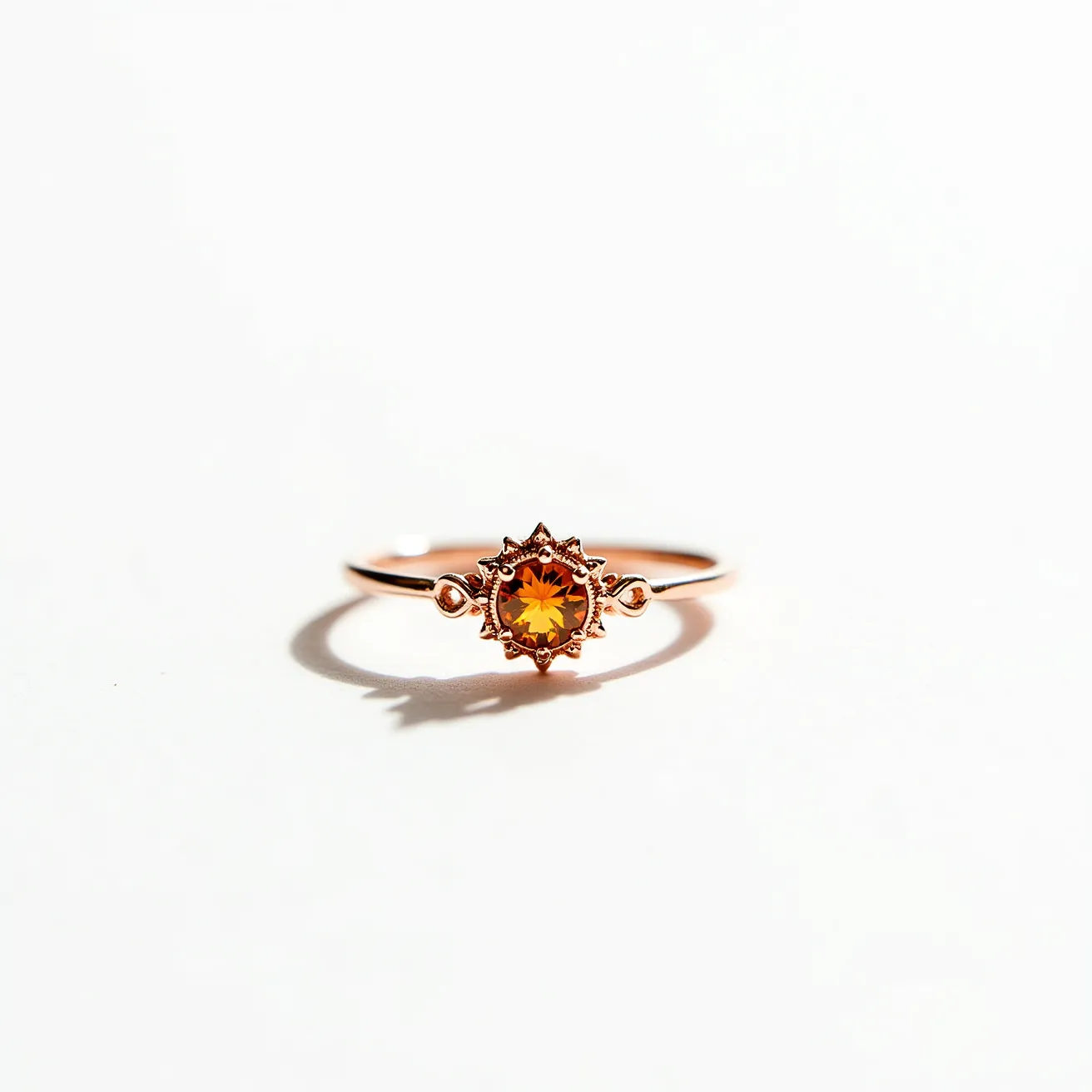 This citrine ring features a round-cut citrine gemstone set into a delicate, ornamental setting crafted from what appears to be rose gold. The citrine, with its warm, golden hue, is the central focus and is securely held in a bezel-like setting that is intricately designed, resembling a floral or sun motif with small petal-like extensions surrounding the stone. The band of the ring is slim and minimalist, designed to complement and emphasize the beauty of the central citrine stone. The ring does not appear to have any additional clasps or attachments, maintaining a simple yet elegant aesthetic.