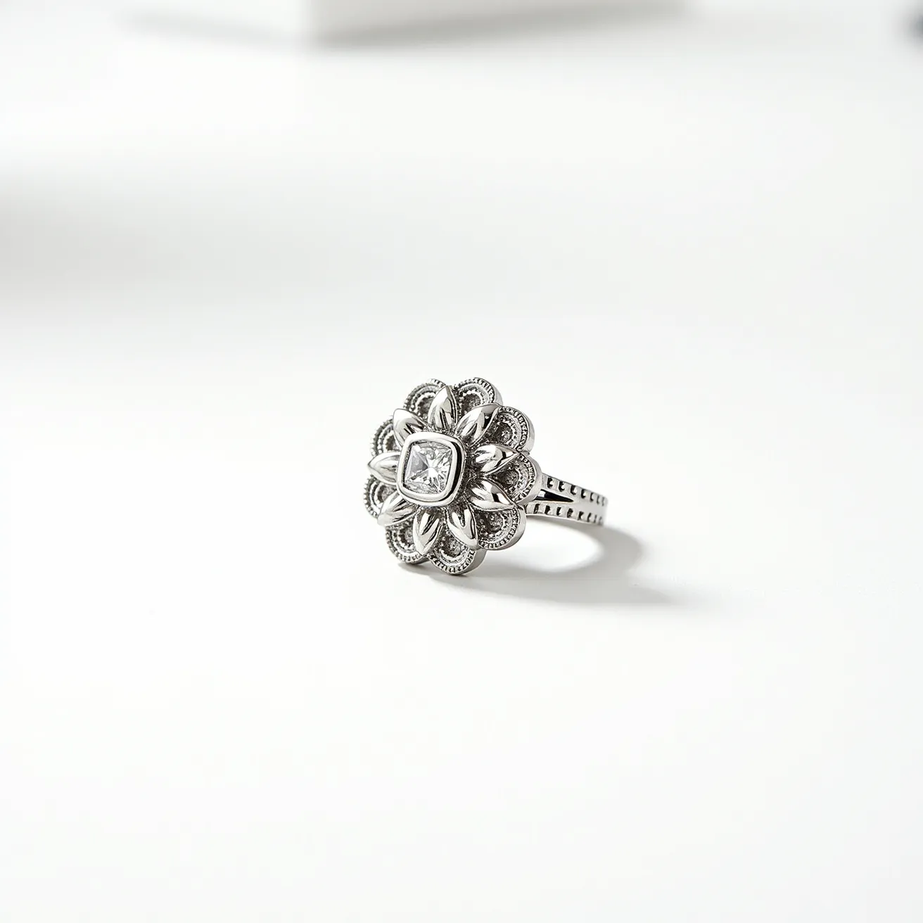 This cocktail ring features a central square-cut gem, possibly a diamond, set in a bezel setting which enhances its elegance and security. The ring is crafted from a white metal, likely platinum or white gold, adding to its sophisticated appeal. Surrounding the central stone is an elaborate floral design made of intricately arranged loops and milgrain detailing, adding texture and depth to the piece. The band of the ring is also adorned with milgrain detailing, creating a cohesive and ornate appearance throughout. The overall design is intricate and suggests a vintage or antique influence, appealing to those who appreciate detailed craftsmanship.