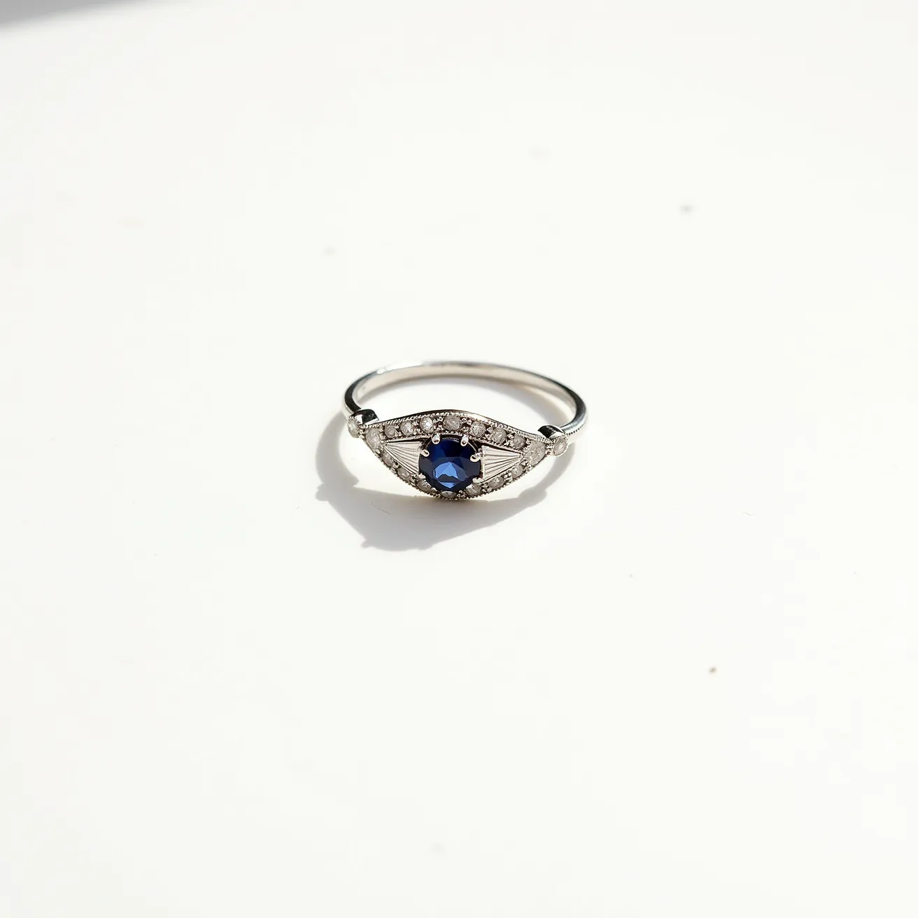 This cocktail ring features a sleek metal band, likely made of white gold or platinum, which enhances its elegant appearance. At its center, a captivating deep blue sapphire takes prominence, cut into a round brilliant shape to maximize its sparkle. The sapphire is set in a secure bezel or prong setting, highlighting its beauty while keeping it firmly in place. Surrounding the sapphire are small, shimmering diamonds or diamond-like stones, arranged in a halo pattern that accentuates the central gem, creating a striking visual effect. The band is simple, ensuring that the focus remains on the intricate design and sparkling gems of the ring's head.