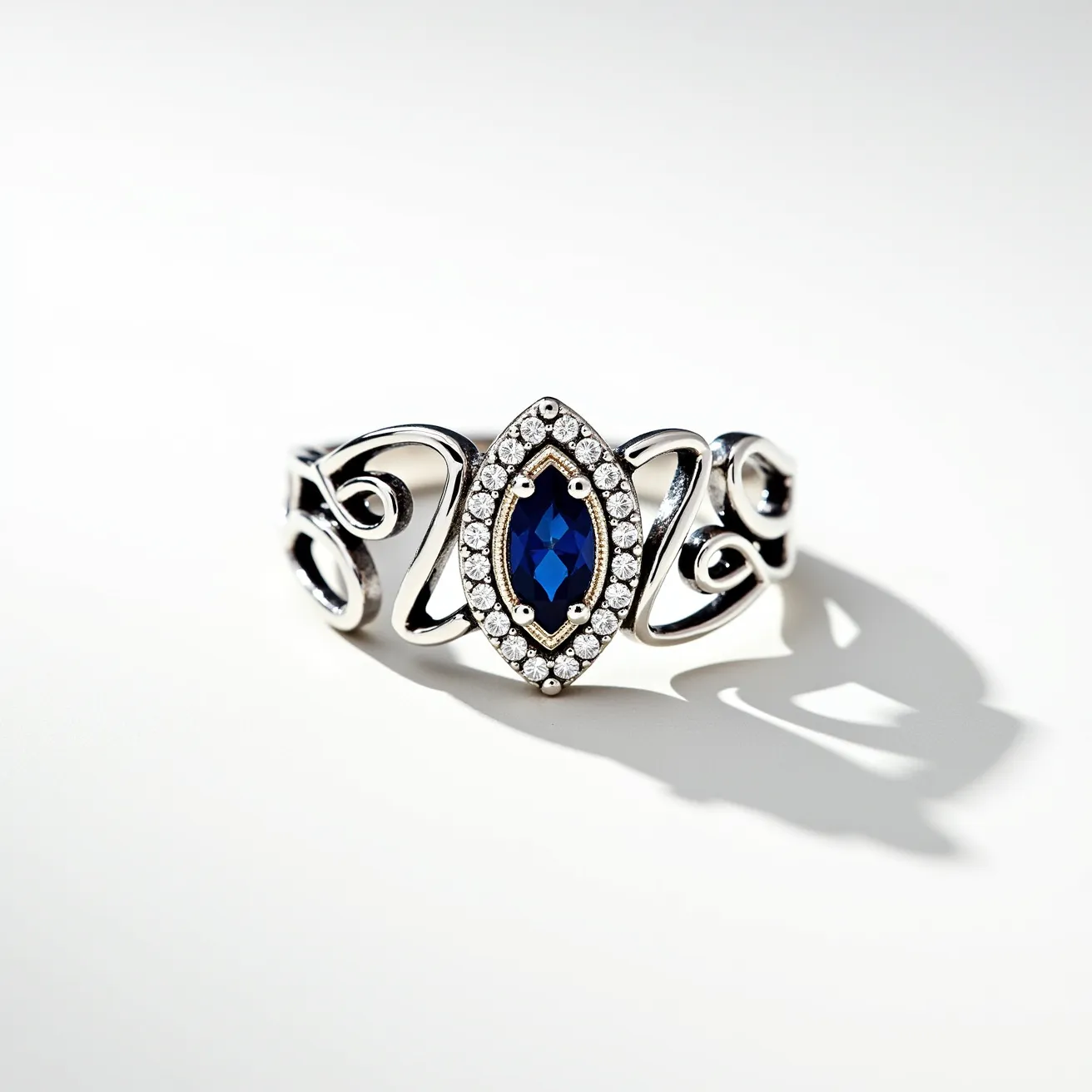 This cocktail ring features a striking marquise-cut blue sapphire at its center, surrounded by a halo of small, sparkling round diamonds set in a delicate pavé setting. The band, crafted from polished silver, showcases an elegant openwork design with flowing scroll motifs on either side of the central stone, enhancing its sophisticated style. There is no visible clasp or attachment, as the ring is designed to be worn as a single, continuous piece. The combination of the deep blue sapphire and bright diamonds creates a stunning contrast that makes this ring an eye-catching statement piece.