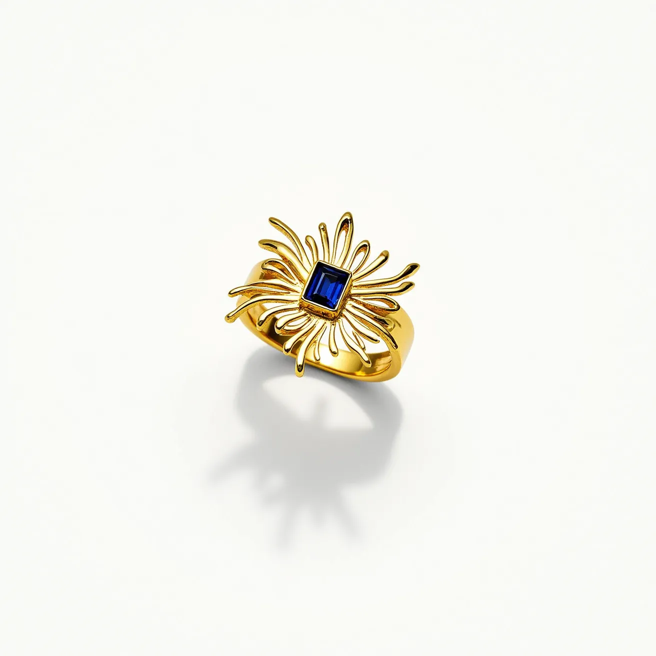 This cocktail ring features a striking design with a central rectangular gem, likely a blue sapphire, cut in an emerald shape. The sapphire is set within an intricate gold framework resembling a burst of rays or petals, giving it a floral or star-like appearance. The setting is elegant, using prongs to enhance the gemstone's visibility and brilliance. The band is made of polished gold, complementing the rich blue of the sapphire and offering a luxurious and bold statement piece. The craftsmanship emphasizes both the beauty of the gem and the artistic flair of the ring’s gold design.