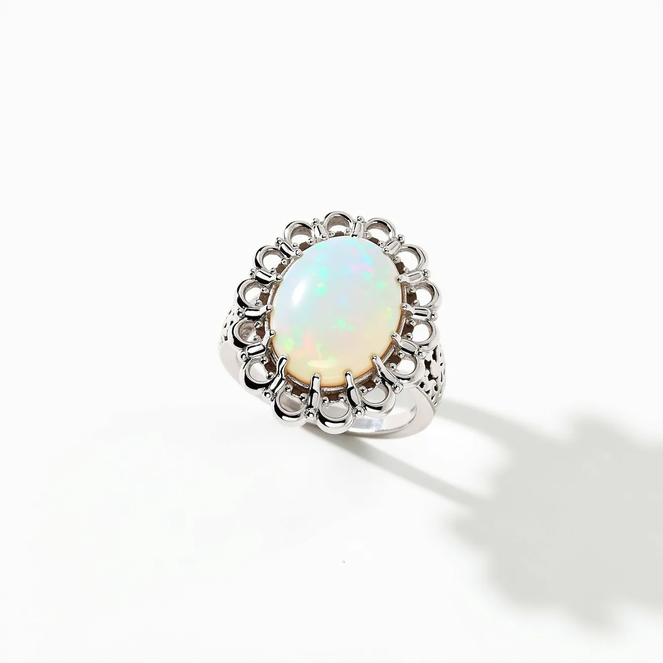 This cocktail ring showcases an exquisite opal gemstone, cut in a smooth oval cabochon style, radiating a captivating array of colors. The opal is set in a prominent prong setting, featuring a metallic base that enhances the stone's vibrant hues. Surrounding the central opal, a decorative halo of metal detailing adds an intricate touch, accentuating the ring's elegant design. The band, crafted from a shiny silver-toned material, merges seamlessly with the ornate setting, offering a harmonious balance between the stone and its setting.