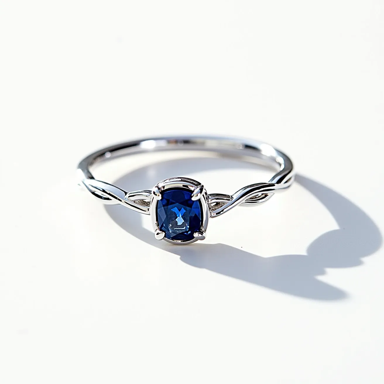 This couple ring features a sleek metal band, likely made of white gold or platinum, which complements the central gemstone. The ring is adorned with a prominent blue sapphire, characterized by its deep hue and cushion cut, providing a sophisticated touch. The sapphire is securely held in place by a four-prong setting, which enhances its brilliance and visibility. The band itself showcases a delicate twist design, adding an element of elegance and uniqueness to the overall aesthetic of the piece.