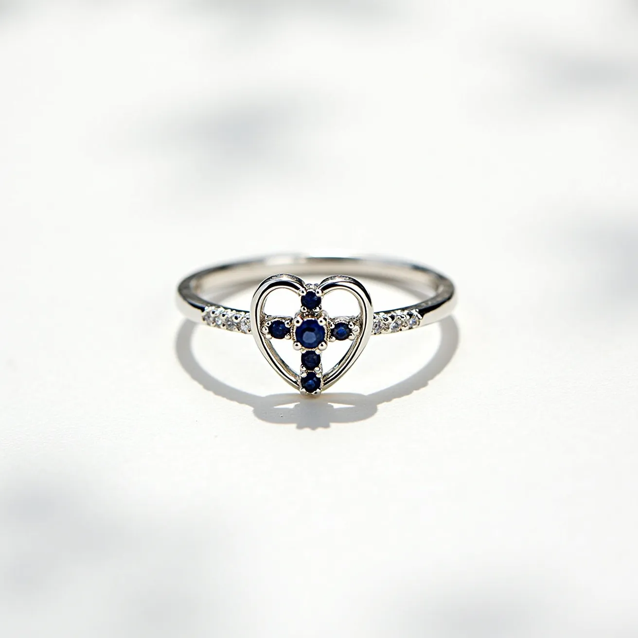 This cross ring features a delicate design composed of a heart shape at the center, crafted from a polished metal that appears to be silver or white gold. The centerpiece showcases a cross motif set with small, round-cut blue sapphires positioned in a prong setting, adding a subtle yet captivating brilliance. The band is adorned with additional small, clear gemstones, likely diamonds or cubic zirconia, also in a prong setting, enhancing the ring's overall sparkle. This elegant piece combines both modern and traditional elements, ideal for those seeking a meaningful yet stylish accessory.