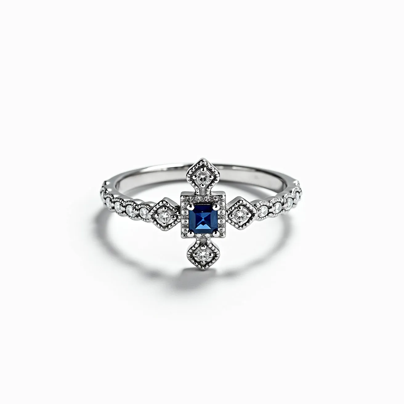 This cross ring features a sleek band crafted from a silver-toned metal, possibly white gold or sterling silver. At its center, a square-cut blue sapphire serves as the focal point, enclosed in a secure bezel setting. The sapphire is elegantly complemented by a series of smaller, round brilliant diamonds arranged in a cross pattern, each set in intricate milgrain creating an antique-inspired look. The ring’s band is further adorned with evenly spaced round diamonds, each held in place by tiny prongs that enhance the ring’s overall sparkle and elegance.