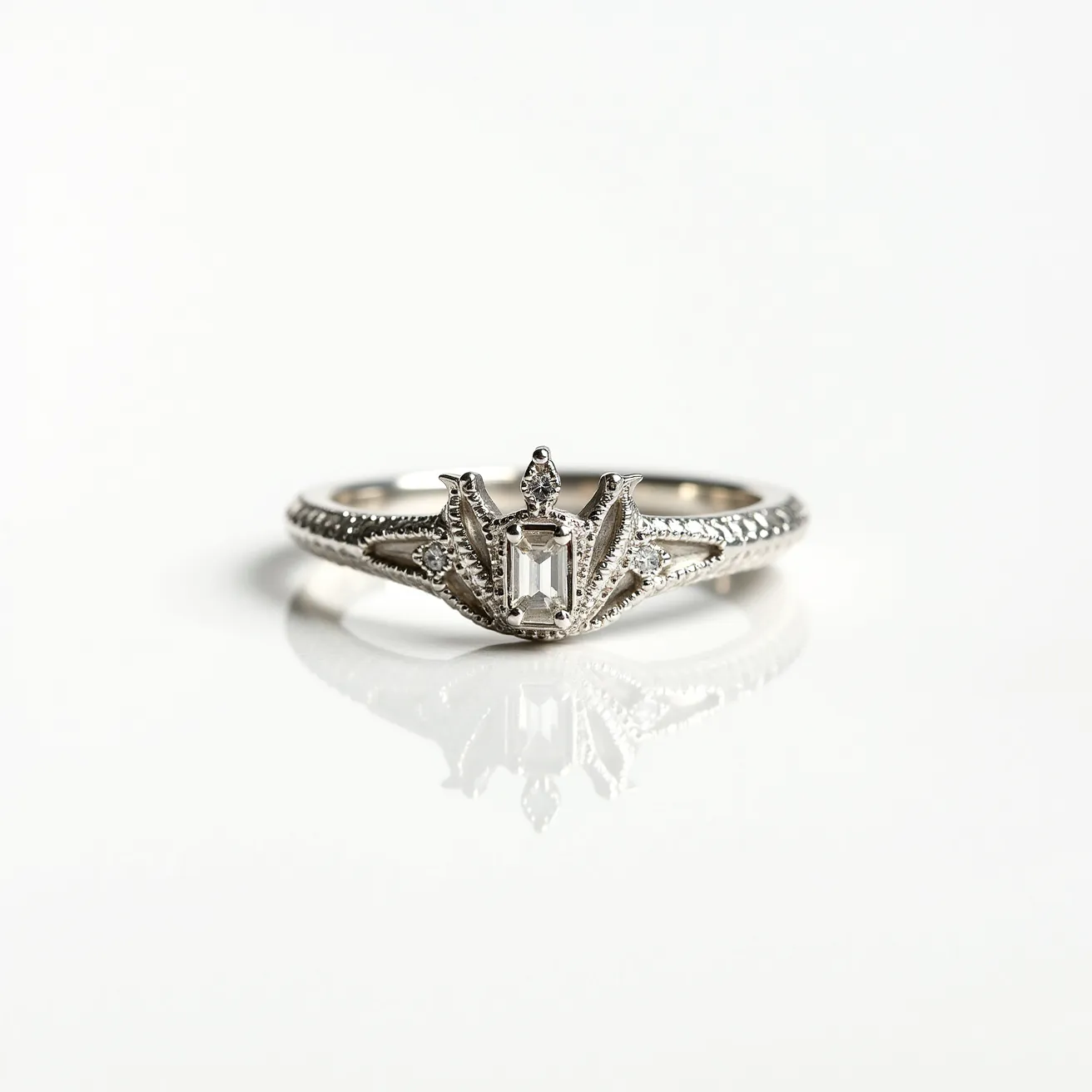 This crown ring features an intricate design crafted from what appears to be a polished silver or white gold metal. The centerpiece of the ring is a baguette-cut gemstone, likely a diamond, set vertically in a bezel setting, creating the shape of a crown. On either side of the central stone, small round-cut diamonds are placed in six-prong settings, adding an elegant sparkle to the overall design. The band itself is adorned with detailed engravings, giving it a textured finish that complements the regal theme. The overall construction showcases skilled craftsmanship with an emphasis on both detail and symmetry.