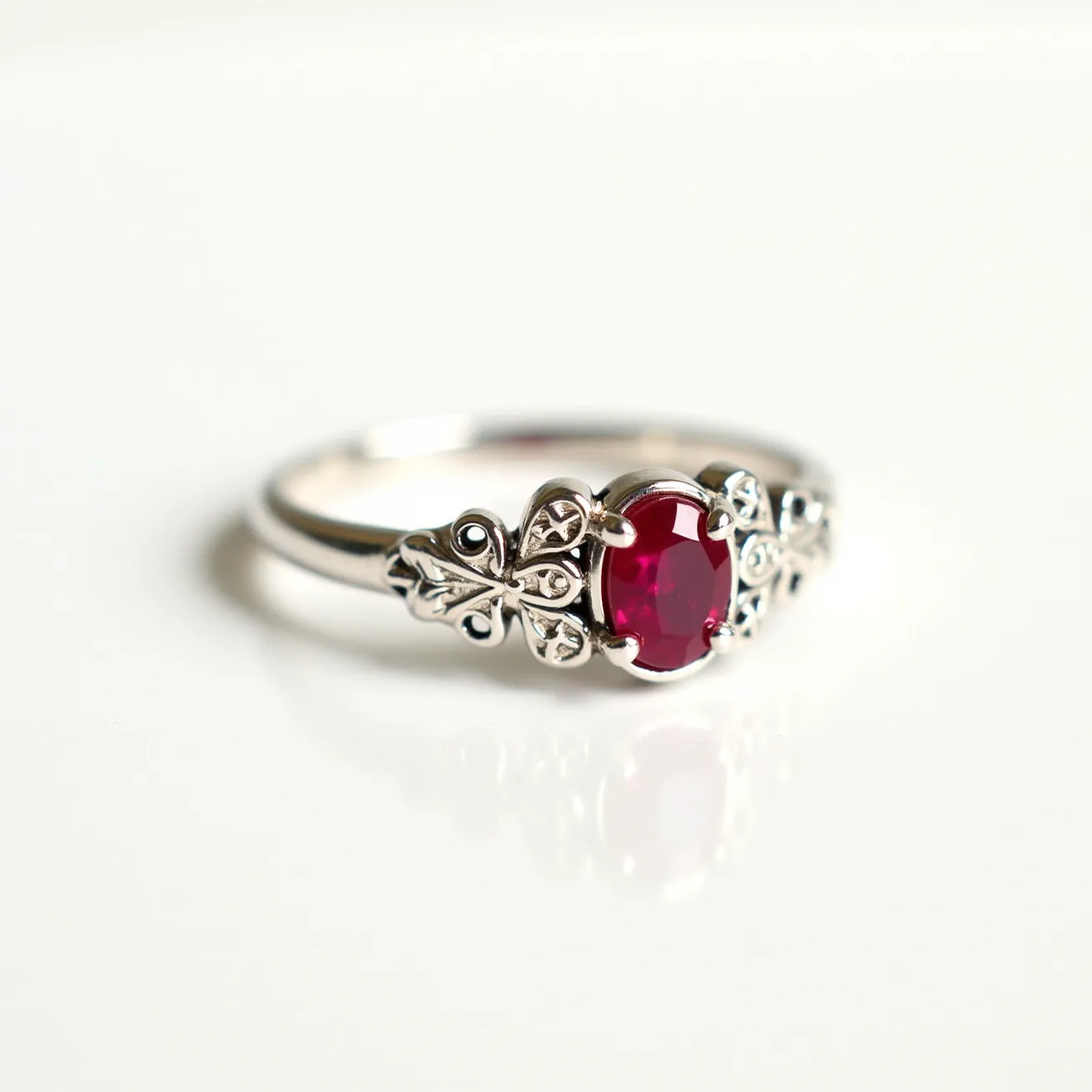 This crown ring features a central oval-cut red gemstone, likely a ruby, set in a well-crafted prong setting. The band appears to be made of a silver metal, enhancing its elegant design. Decorative elements resembling fleur-de-lis motifs adorn the ring on either side of the gemstone, adding to its regal aesthetic. The ring's overall design is intricate, and the craftsmanship emphasizes the central gemstone beautifully, making it a striking piece of jewelry.