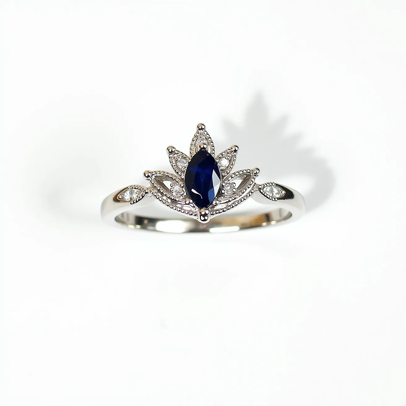 This crown ring features a central marquise-cut blue sapphire as the focal gemstone, elegantly set within a prong setting to accentuate its deep hue and elongated shape. Surrounding the sapphire are intricate details reminiscent of a crown, embellished with small round-cut diamonds that add brilliance through their pavé setting. The band appears to be made of white gold, offering a sleek and polished finish that complements the overall design. The delicately crafted elements of the ring highlight its regal inspiration, creating a sophisticated piece of jewelry with both elegance and sparkle.