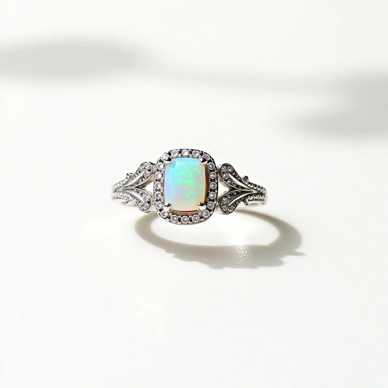 This custom ring features a stunning central opal gemstone, cut into an elegant cushion shape. The opal is set in a halo setting adorned with round brilliant-cut diamonds that enhance its iridescent play of color. The band is crafted from a material that appears to be silver or white gold, intricately designed with detailed embellishments flanking the center stone. The delicate band further includes additional small diamonds, contributing to the overall sparkle and elegance of the piece. The craftsmanship and choice of materials make this ring a unique and exquisite piece of jewelry.