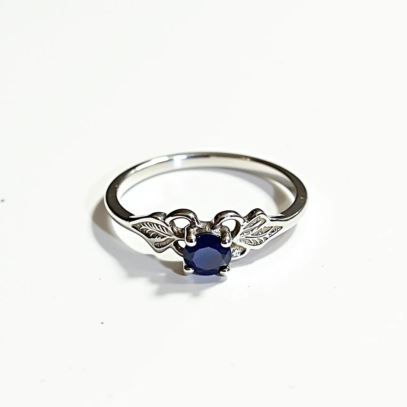 This custom ring features a sleek, polished band crafted from a silver-toned metal, likely sterling silver. At its center is a round, deep blue stone resembling a sapphire, securely held in place by a four-prong setting. The ring's design includes two intricately detailed leaf motifs flanking the central stone, adding an elegant, nature-inspired touch. The combination of materials and details highlights the craftsmanship and aesthetic appeal of this unique piece.