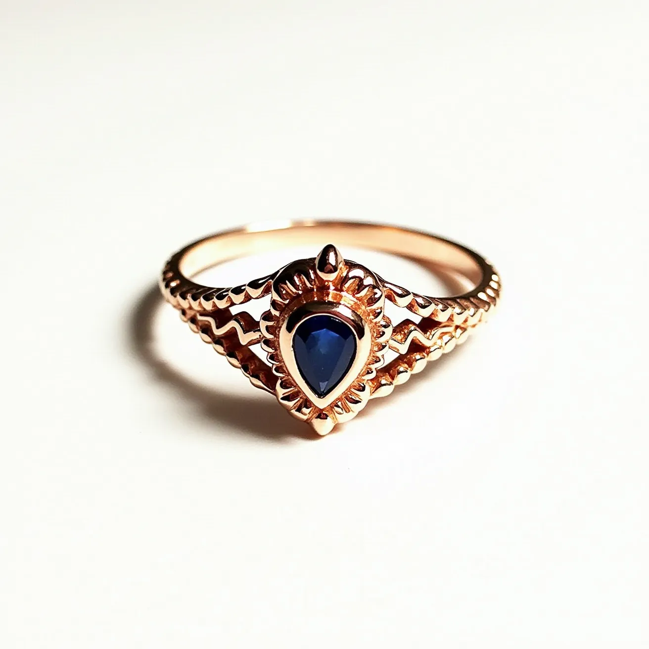 This custom ring features an intricately designed band made of what appears to be rose gold, characterized by a series of smooth scalloped patterns along its side. At its center, the ring showcases a pear-shaped blue gem, likely a sapphire, which is held securely in a bezel setting that ensures its prominence and stability. The bezel setting is adorned with a petal-like design, adding a decorative touch that enhances the overall elegance of the piece. The combination of the deep blue gem with the warm hue of the metal creates a striking contrast.