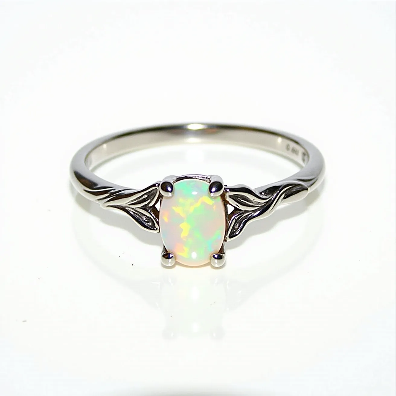 This custom ring features a central oval opal, set in a prong setting, which beautifully showcases the stone’s vibrant play of color. The band is crafted from polished metal, likely silver or white gold, and displays intricate leaf motifs on either side of the gemstone, adding an elegant and organic touch to the design. The opal's iridescence is complemented by the simplicity and elegance of the band, bringing out the luster and uniqueness of the center stone.