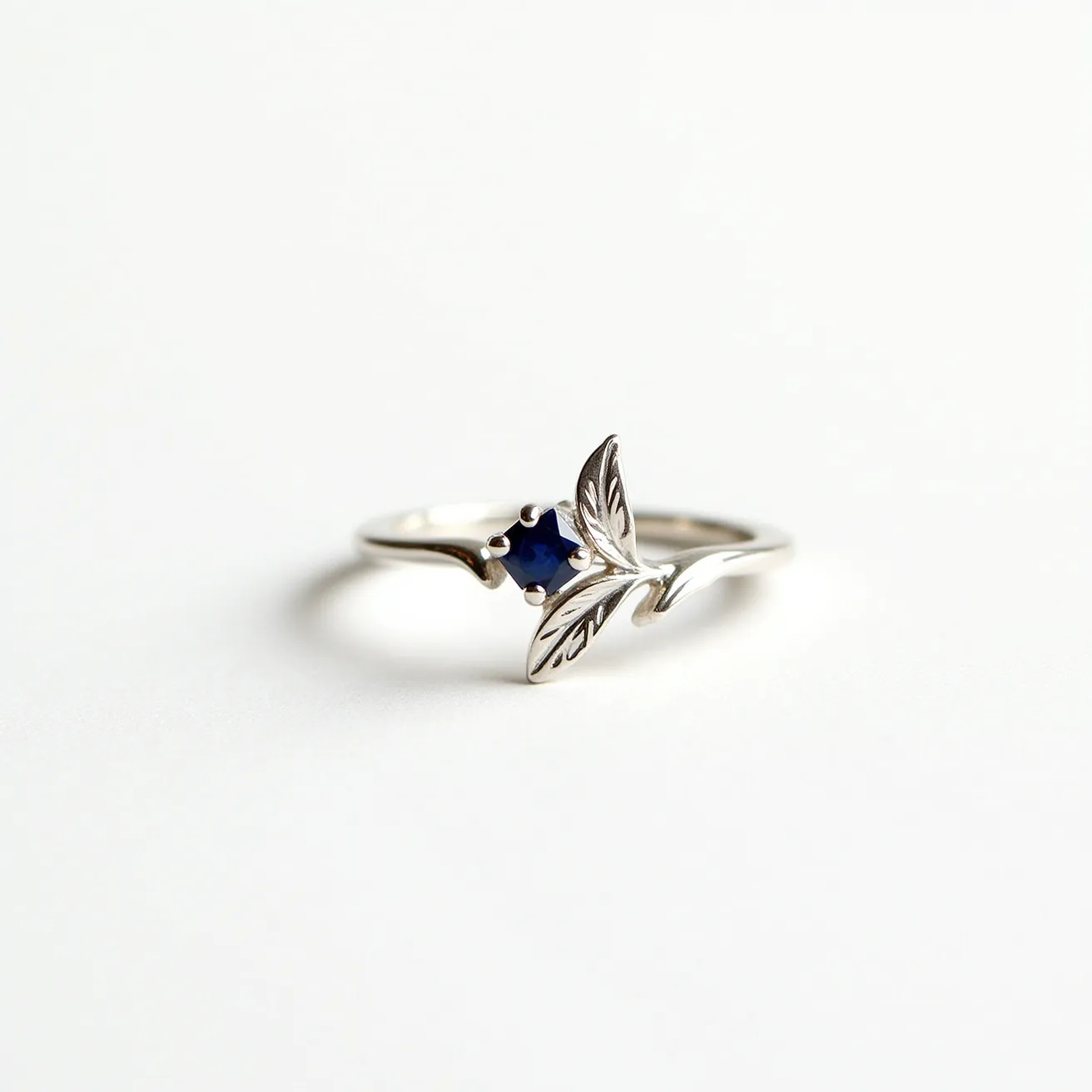 This custom ring features a delicate and intricate design crafted from a silver-toned metal. At its center, a single blue sapphire gemstone, cut in a round faceted style, is set in a prong setting, which securely holds the stone and allows light to enhance its vibrant color. The ring's band is adorned with a unique leaf motif, with detailed engravings that add elegance and a nature-inspired aesthetic. The overall design combines simplicity and sophistication, making it a distinctive piece.