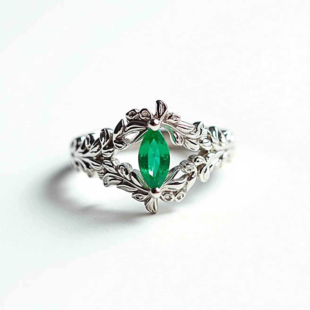 This custom ring features an intricate design crafted from silver-toned metal, adorned with elaborate leaf motifs that create a nature-inspired look. At the center, there is a striking marquise-cut green gem, securely set within the leaf design, enhancing its elegance and prominence. The gem is held in place with multiple prongs that blend seamlessly with the metalwork, ensuring both security and aesthetic coherence. The detailed craftsmanship and natural theme make this ring a unique and captivating piece.