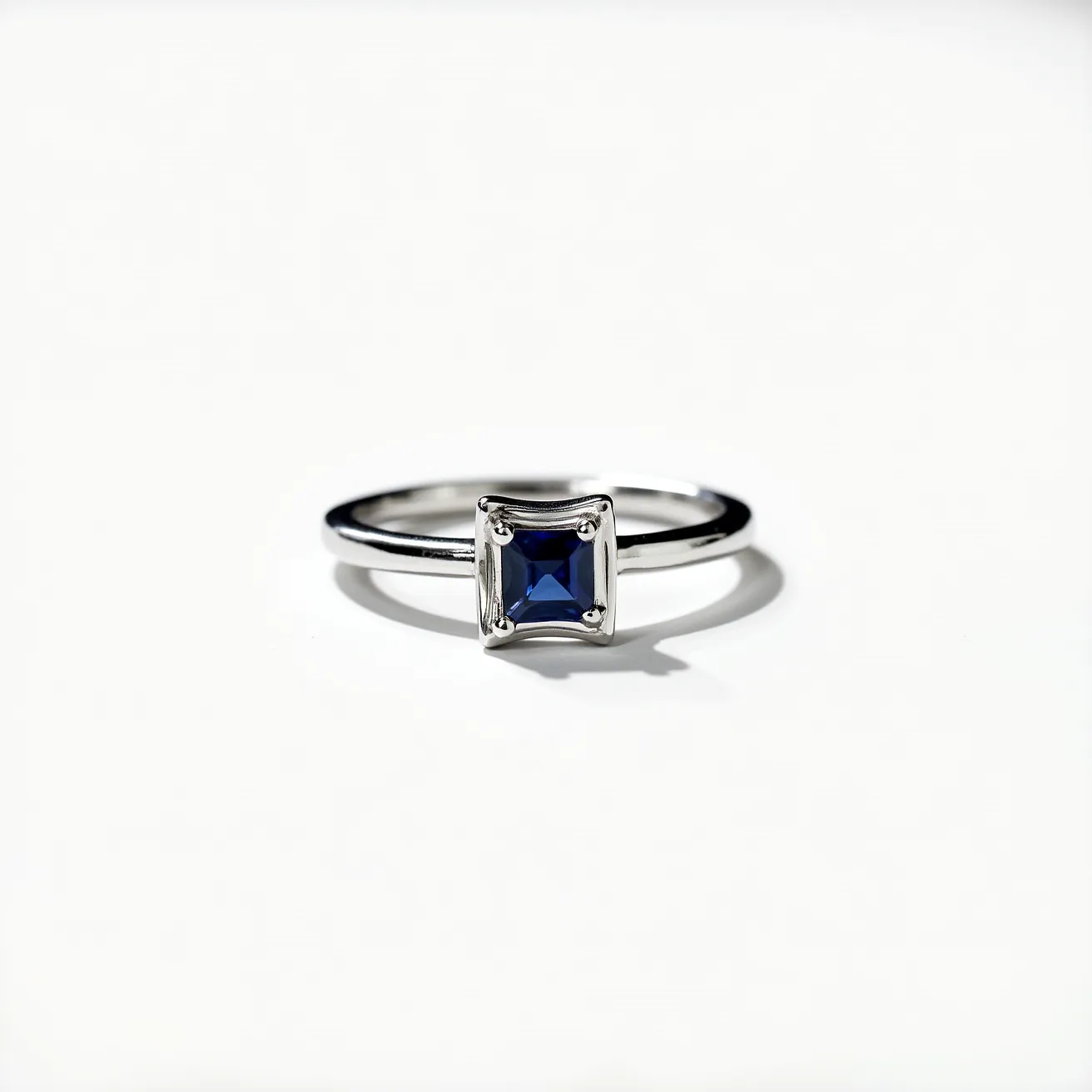 This custom ring features a sleek band crafted from polished metal, possibly white gold or platinum, lending it a modern and elegant appearance. At the center, a striking blue gemstone, shaped in a princess cut, is securely set in a bezel setting, which provides both protection and a contemporary look. The stone's deep blue hue and precision cut enhance its brilliance and elegance, making it the focal point of the design. There are no additional clasps or attachments, allowing the simple elegance of the gemstone and band to shine as the stars of this custom piece.