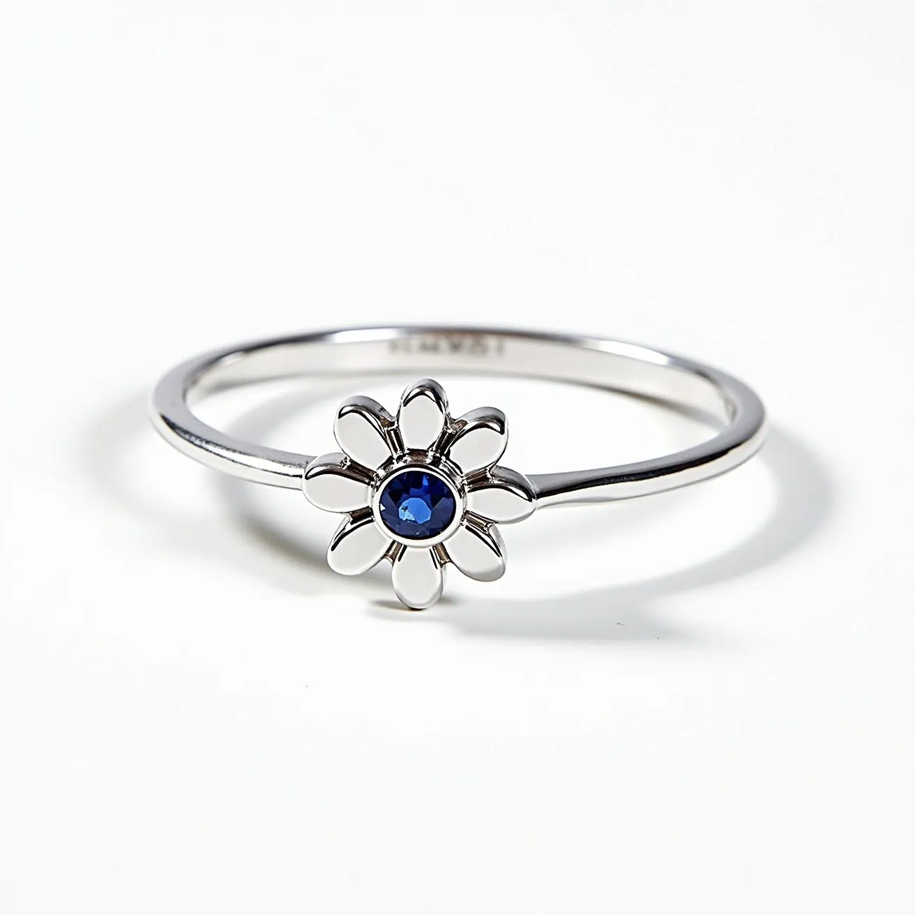 This daisy ring features a delicate design with a band likely crafted from silver or white gold, enhancing its elegant and polished appearance. At the center of the daisy motif is a round-cut blue gemstone, possibly a sapphire, held securely in a bezel setting, which adds a vibrant splash of color to the piece. The daisy petals are crafted with smooth, rounded edges, complementing the central gem. The band is slender and simple, allowing the floral centerpiece to stand out as the focal point.