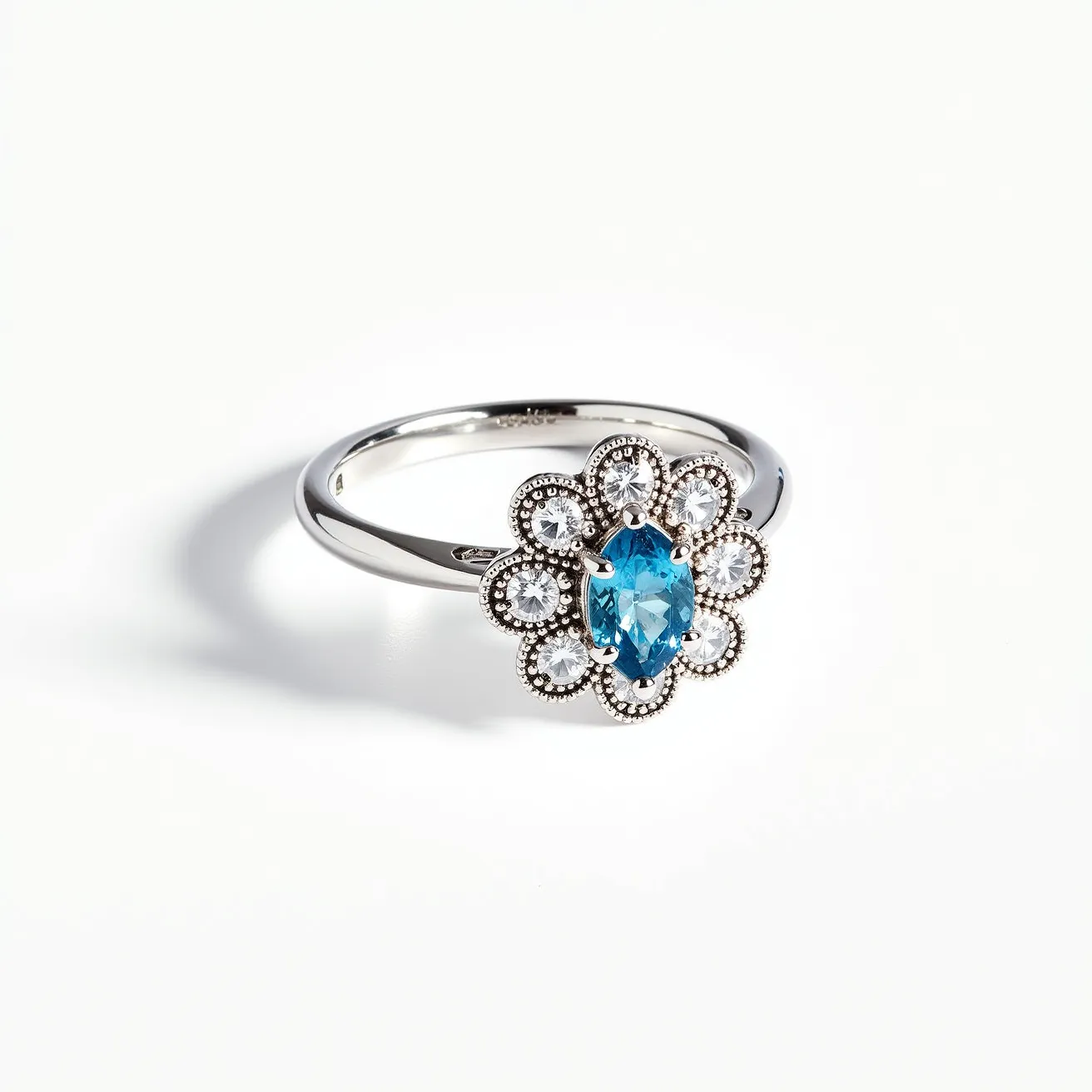 This daisy ring features a striking design with a central oval-cut blue gemstone set in a prong setting, complemented by a surrounding halo of smaller round clear stones. The arrangement creates a floral motif reminiscent of a daisy. The band appears to be made of a shiny metal, possibly white gold or platinum, enhancing the elegance of the piece. The ring showcases intricate detailing around the halo, adding to its vintage charm without any visible clasps or attachments.