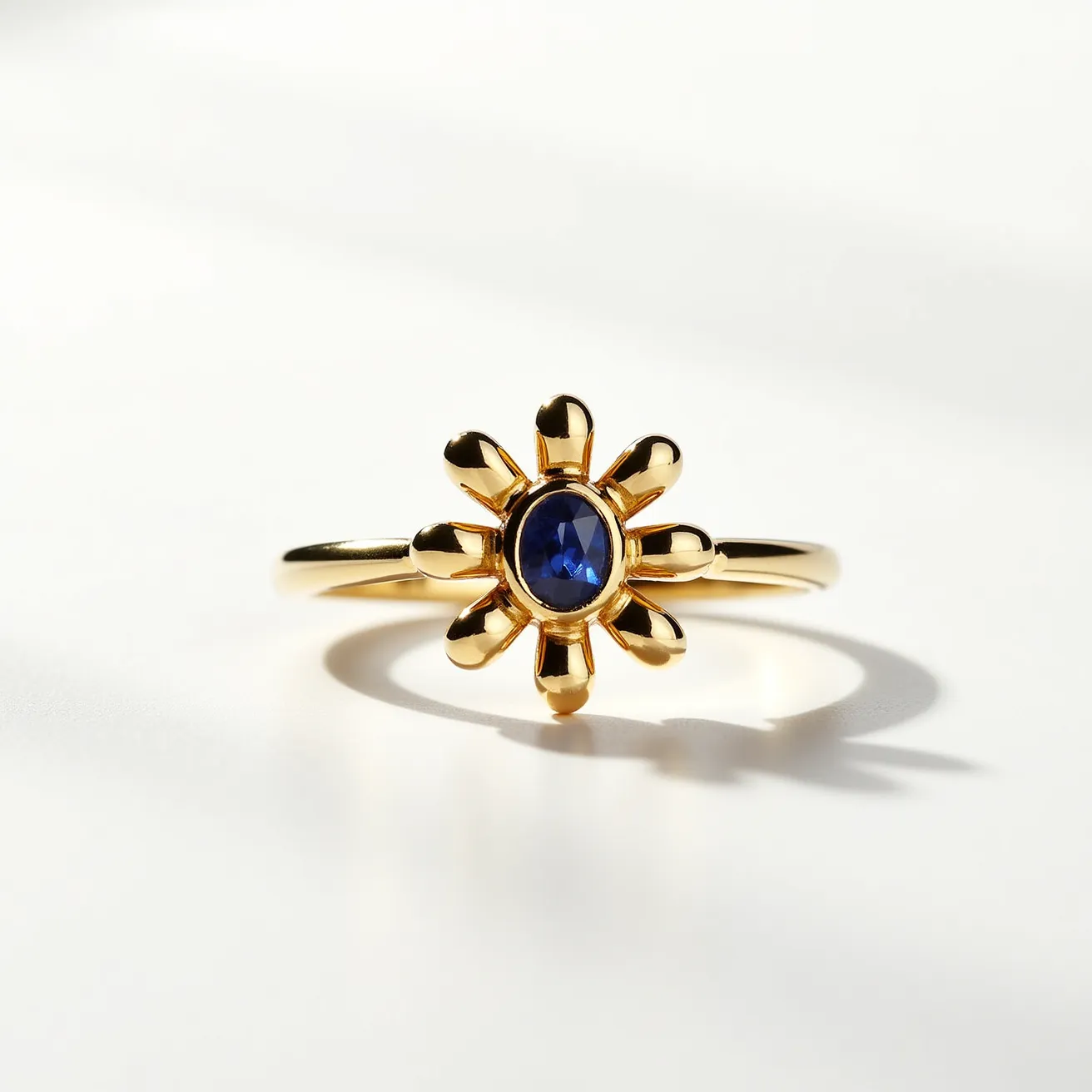 This daisy ring features a delicate design with a central blue gemstone, likely a sapphire, in a round cut, set prominently in a bezel setting. Surrounding the gemstone are petal-like protrusions that form the shape of a daisy, crafted from a gold-colored metal that gives the piece a luxurious appearance. The gold band adds elegance and simplicity, allowing the daisy motif to be the focal point. The ring appears seamless, with no visible clasp or attachment, enhancing the fluidity and continuity of the design.