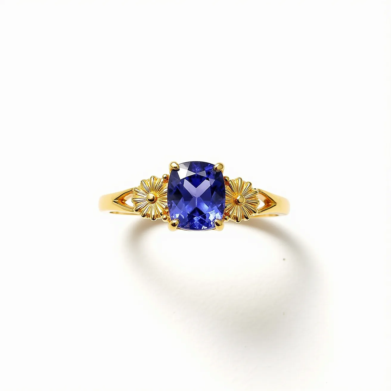 This December birthstone ring showcases a captivating blue stone, likely a tanzanite or blue topaz, cut in a cushion shape. The gemstone is elegantly set in a four-prong setting, which secures it while allowing maximum light to enhance its brilliance. The band is crafted from a yellow gold material, accentuating the deep blue hue of the central stone. On either side of the gemstone, there are intricate floral embellishments, adding a touch of elegance and sophistication to the design. The combination of the blue gemstone with the gold band and detailed floral accents results in a stunning and elegant piece.