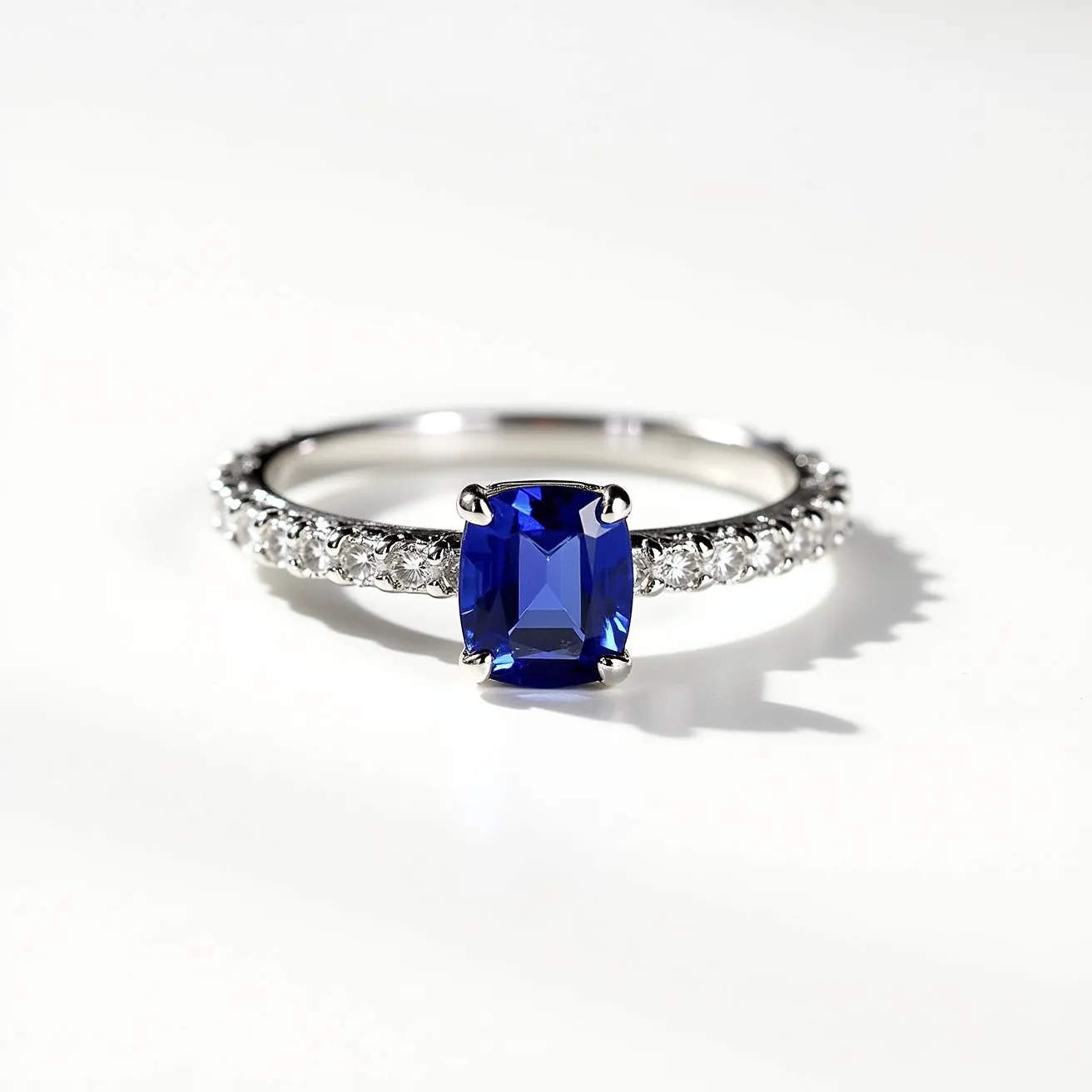 This December birthstone ring features a striking blue gemstone, likely a blue topaz or tanzanite, which is common for December. The gem is cushion-cut and set in a four-prong setting, allowing the stone to catch the light beautifully. The band is adorned with small, round-cut diamonds set in a pave style, adding a touch of sparkle and enhancing the overall elegance of the piece. The band appears to be crafted from a polished white metal, possibly white gold or platinum, complementing the cool tones of the gemstones. There is no visible clasp or attachment as the design features a continuous band typical of a ring.