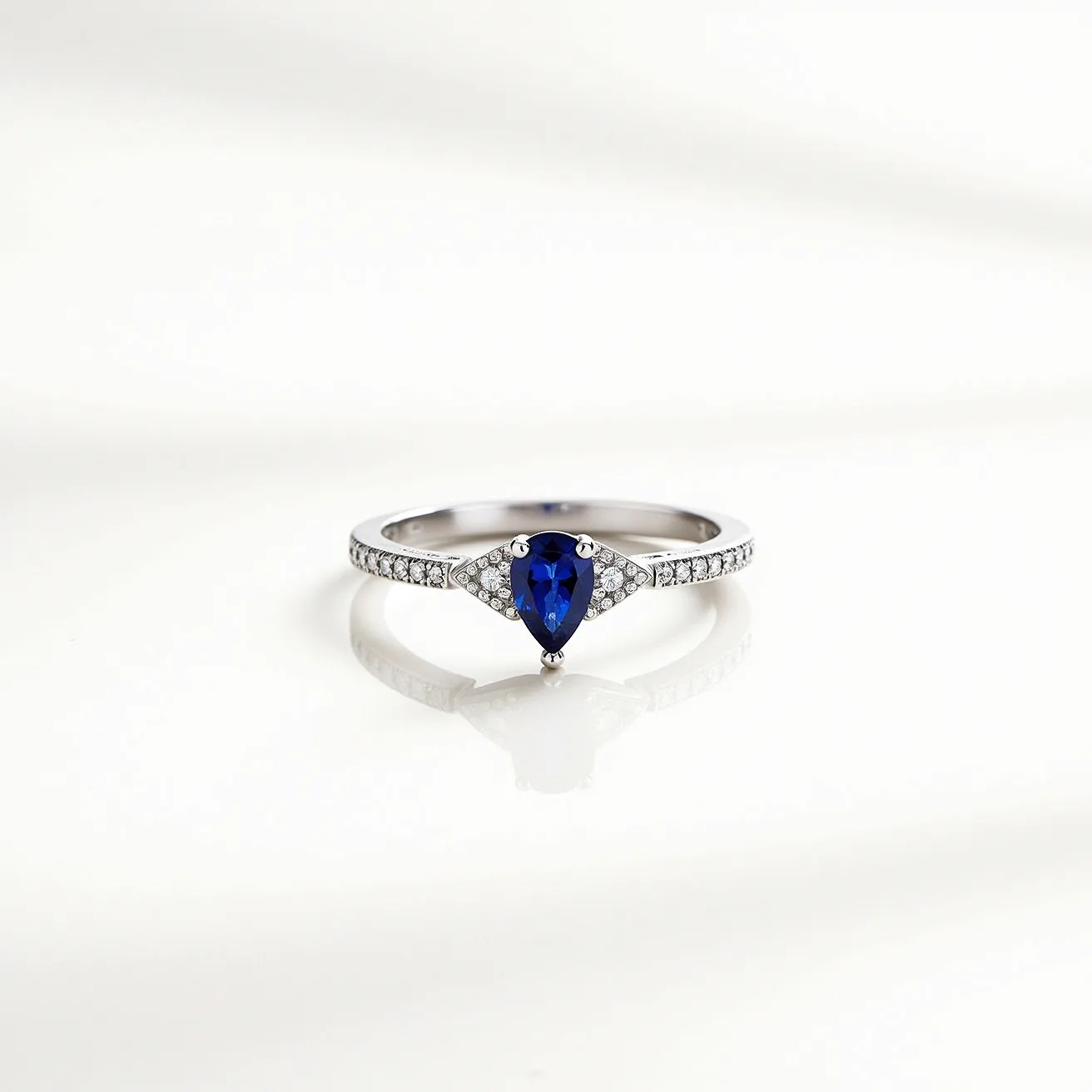 This December birthstone ring features a striking pear-shaped blue stone, likely blue zircon or tanzanite, set in a prong setting that accentuates its vibrant hue. Flanking the central gem, a series of small, round-cut diamonds are delicately embedded in the band, which is composed of a polished white metal, possibly white gold or platinum, providing a sleek and elegant appearance. The diamonds on the band enhance the ring's brilliance, making it a sophisticated piece that represents December's birthstone beautifully.