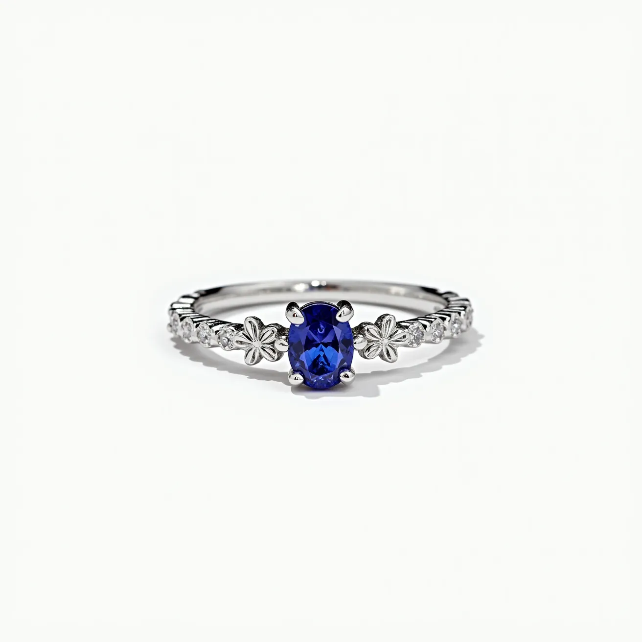 This December birthstone ring features a stunning oval-cut blue tanzanite, securely held in place by a four-prong setting. The ring is crafted from polished white metal, likely silver or white gold, which complements the vibrant hue of the central gemstone. The band is intricately designed with alternating small flowers and circular settings, each housing a sparkling white accent stone, creating a harmonious and delicate adornment. The combination of the floral motifs and accent stones adds elegance and intricacy to the overall design.