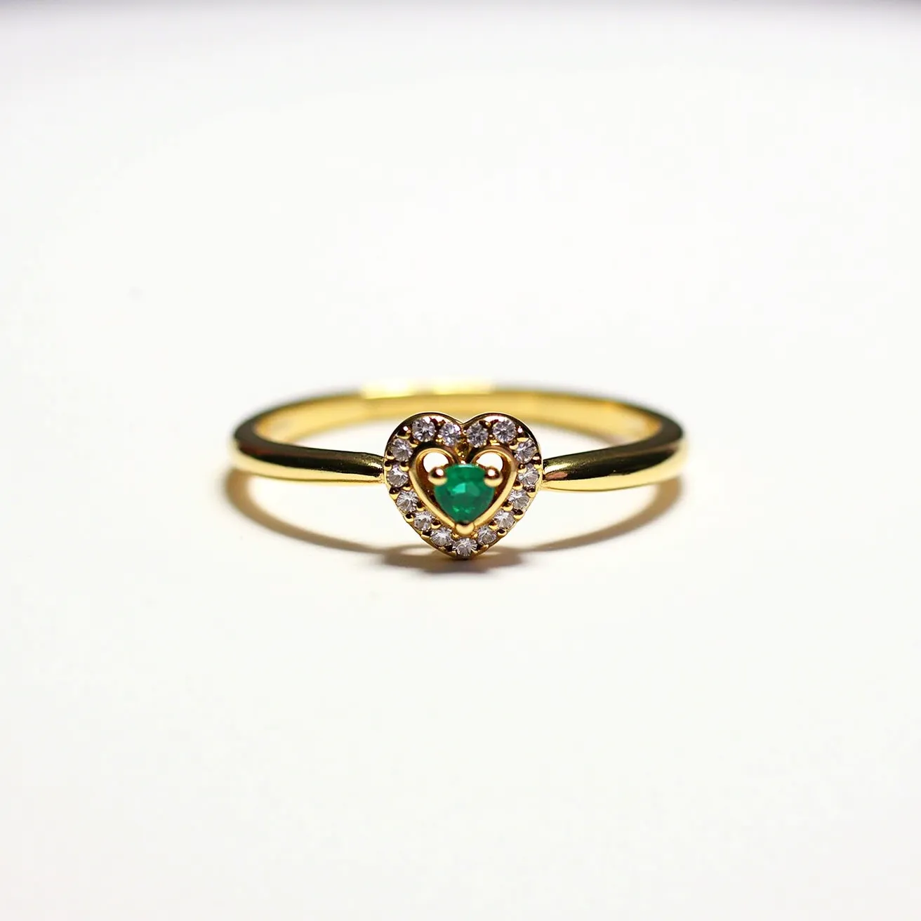 This diamond heart ring features a central emerald stone, round in shape, showcased in a heart-shaped setting. The emerald is surrounded by a halo of small, round brilliant-cut diamonds, enhancing the sparkle and elegance of the piece. The band, crafted from yellow gold, complements the brilliance of the emerald and diamonds. The setting securely holds the stones without visible clasps or attachments, allowing the heart shape to stand out prominently. The combination of precious stones and metal creates a timeless and classic design perfect for romantic occasions.