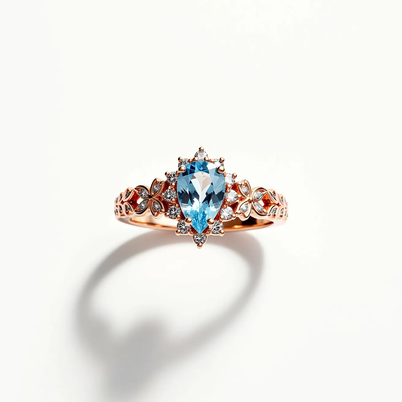 This diamond ring features a prominent pear-shaped blue gemstone seated at its center, likely aquamarine, accented by a halo of small round diamonds. The ring band is crafted from a rose gold material with intricate detailing, including floral motifs that add an elegant touch to its design. The diamonds are prong-set, which enhances their brilliance and securely holds them in place, complementing the central stone beautifully. The floral elements are seamlessly integrated into the band, contributing to the overall aesthetic and making it a captivating piece of jewelry.