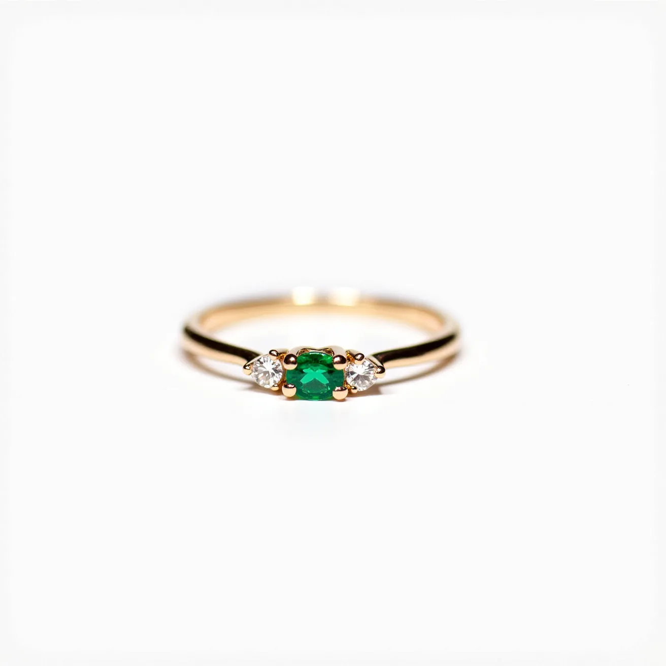 This diamond ring features a central emerald stone, cut in an oval shape and set in a four-prong setting. Flanking the emerald are two smaller, round-cut diamonds, each held in a prong setting on either side. The band appears to be crafted from yellow gold, providing a classic and elegant contrast to the vibrant green of the emerald and the sparkle of the diamonds. The combination of these elements highlights the ring's sophisticated and timeless design.