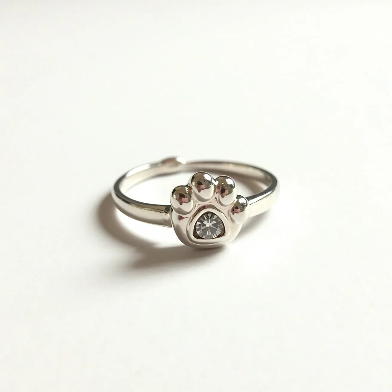 This dog ring features a sleek silver band crafted into an adjustable design, with a charming paw motif as its focal point. At the center of the paw, a small, round-cut gemstone is securely set, adding a subtle sparkle to the piece. The simplicity of the design, coupled with the polished finish of the metal, creates an elegant yet playful accessory that celebrates a love for dogs. The band appears to have an open-ended style allowing for easy size adjustments, making it a versatile addition to any jewelry collection.