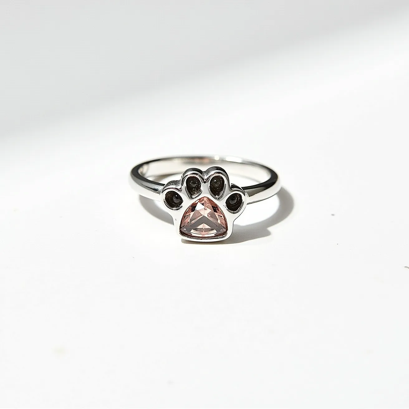 This dog ring features a stylish design with a silver band that showcases a paw print motif. At the center of the paw print, a triangular-cut pink stone is set, giving the ring a touch of elegance. Each toe of the paw is detailed with a small, dark setting, possibly containing tiny gemstones or enamel. The overall look is modern and chic, making it an eye-catching accessory. There is no visible clasp or attachment, indicating it is a standard band style ring.