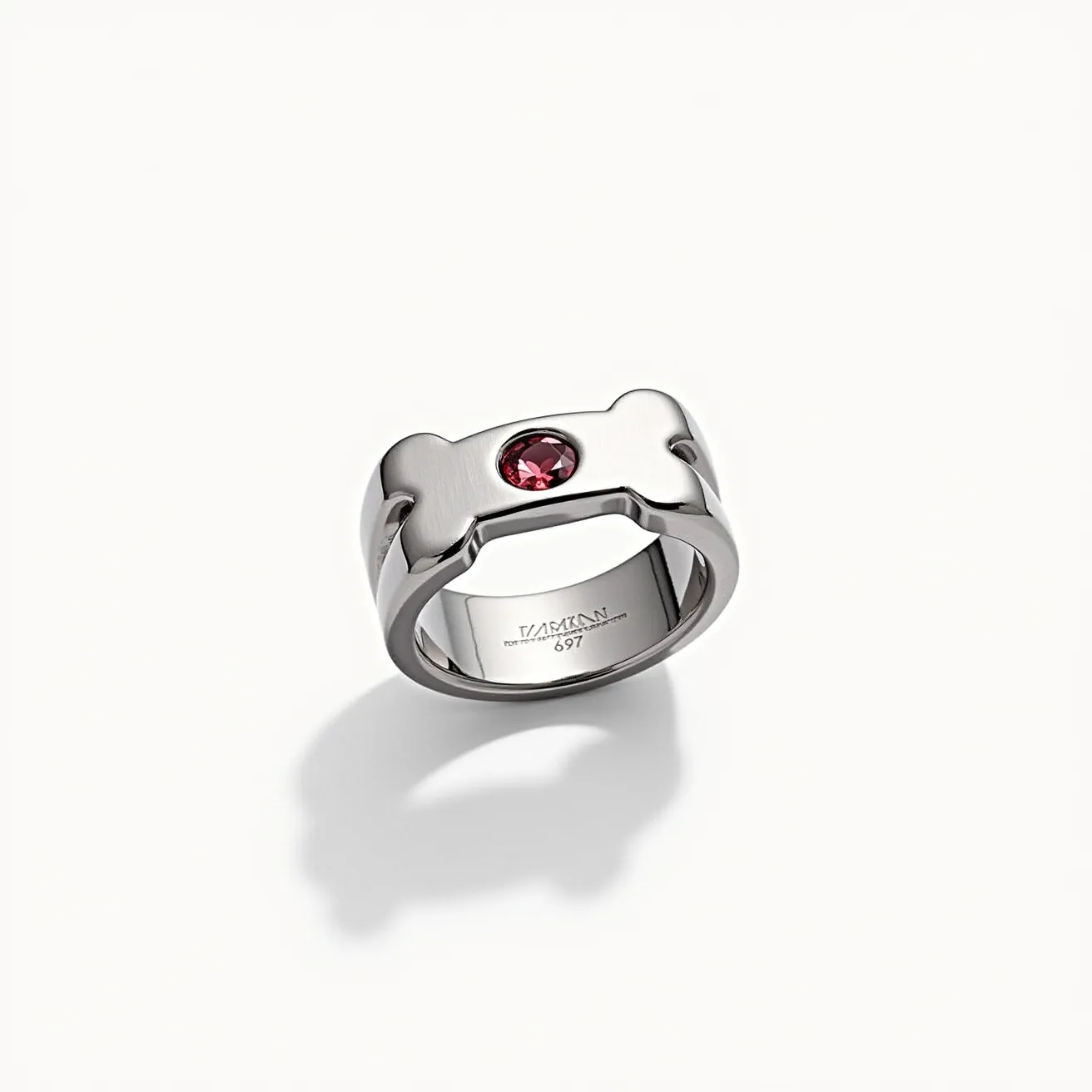 This dog ring features a sleek metallic construction, possibly stainless steel or silver, with a distinct dog bone shape integrated into its design. At the center, a round-cut gemstone, likely a deep red ruby, is securely set, enhancing the piece with a touch of elegance. The stone is bezel-set, emphasizing a modern and refined aesthetic. The interior of the ring is smooth and polished, ensuring comfort for the wearer. An inscription inside the band adds a subtle, personalized detail to this unique piece of jewelry.