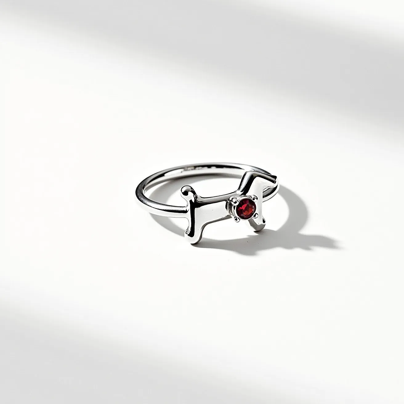 This dog ring is crafted from a polished metallic material, likely silver in appearance, shaped into a stylized outline of a dog. At the center of the dog's body, there is a round, faceted red gemstone set in a simple bezel setting, adding a touch of color and elegance to the piece. The band is smooth and minimalistic, blending seamlessly into the outline of the dog, with no additional clasps or attachments visible.