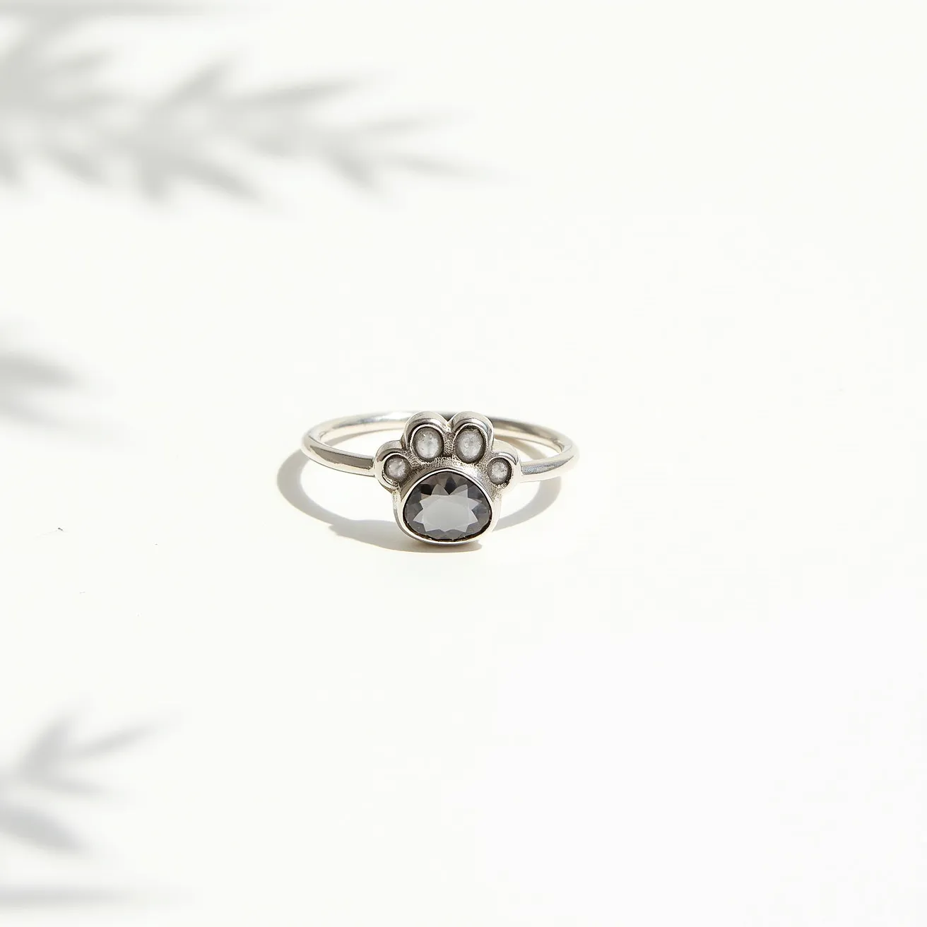 This dog ring features a delicate silver band with a paw-shaped design as its centerpiece. The paw includes an oval-cut dark gemstone set in a bezel setting, which forms the larger pad of the paw. Surrounding the central stone are smaller, round silver elements representing the toe pads. The simplicity of the ring's design highlights the elegant contrast between the silver and the dark gemstone, making it a charming piece of jewelry for dog lovers.