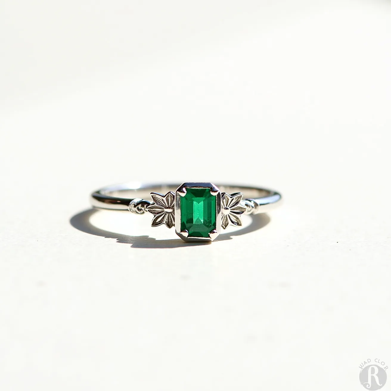 This emerald birthstone ring features a vibrant, rectangular emerald-cut stone set in a sleek, silver band. The gem is elegantly held by a prong setting that ensures its security while enhancing its brilliance. The band is adorned with intricate leaf-shaped accents on either side of the stone, adding a touch of nature-inspired elegance. The overall design balances simplicity and sophistication, ideal for those who appreciate classic beauty with a hint of decorative detail.