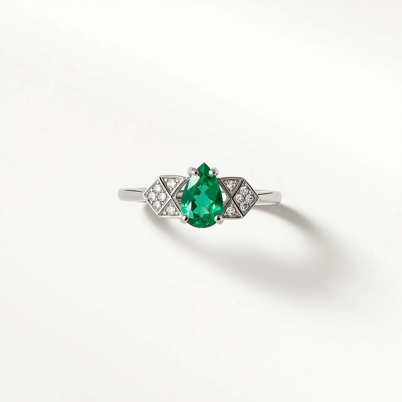 This emerald birthstone ring features a striking pear-cut emerald set prominently at its center. The emerald is brilliantly framed by numerous small round diamonds, intricately set into a geometric configuration on either side, accentuating the vibrant green hue of the main stone. The ring's band is crafted from a polished metal, likely platinum or white gold, providing a sleek and elegant backdrop that enhances the overall sparkle of the stones. The setting secures the stones with prongs, ensuring both security and optimal light reflection. The combination of emerald and diamonds offers a classic yet modern aesthetic that makes this ring a timeless piece of jewelry.