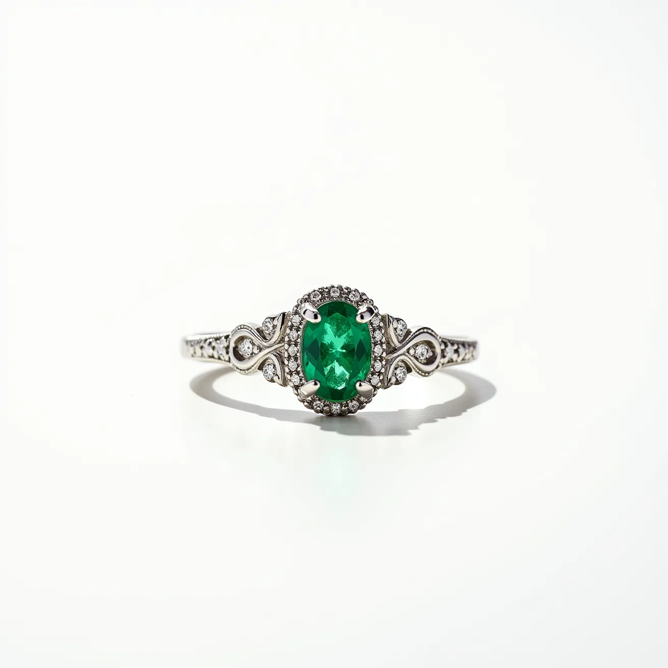 This emerald birthstone ring features a vibrant oval-cut emerald as the centerpiece, beautifully set in a four-prong setting. The emerald is surrounded by a halo of small, round diamonds enhancing its striking color and adding extra sparkle to the design. The band is crafted from a lustrous white metal, possibly white gold or platinum, and is adorned with intricate scrollwork and additional small diamonds along the sides, adding elegance to the ring. The craftsmanship emphasizes the regal allure of the emerald, making it a stunning piece for any jewelry collection.