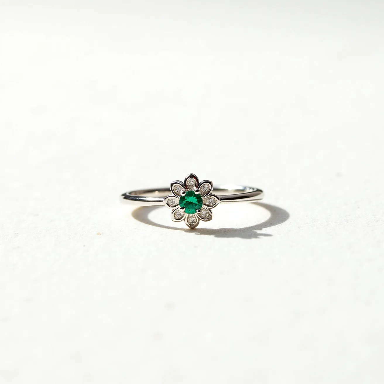 This emerald birthstone ring is elegantly crafted with a delicate band likely made of a polished metal such as white gold or platinum. At its center, the ring features a vibrant, round-cut emerald, which is surrounded by a floral arrangement of small, clear stones resembling diamonds. These surrounding stones are set in a petal-like pattern that enhances the central emerald's striking color. The overall design blends simplicity with a touch of intricate detailing, giving it a refined and timeless appeal.