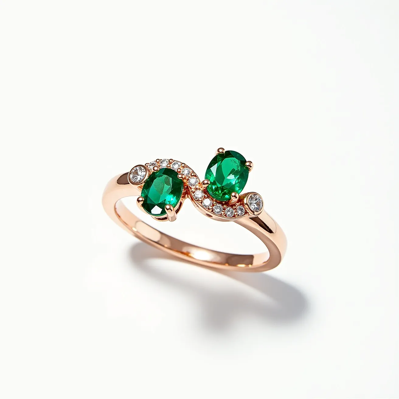 This emerald engagement ring features two stunning oval-cut emerald gemstones set in a graceful rose gold band. The emeralds are prominently displayed in prong settings, which enhance their rich green brilliance. Surrounding the emeralds, a delicate array of small round-cut diamonds forms an elegant swirl pattern, adding sparkle and sophistication. On each side of the swirl, additional round diamonds are embedded, providing a balanced and symmetrical design. The band itself is sleek and polished, completing the luxurious aesthetic with a modern yet timeless appeal.