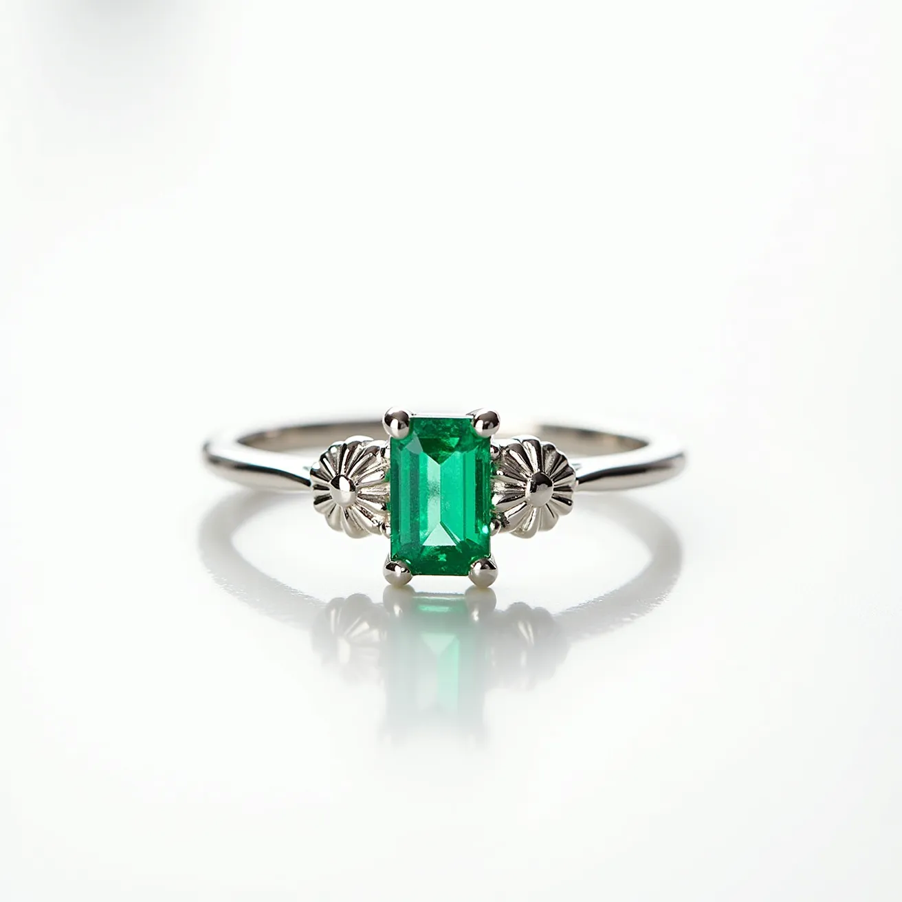 This emerald promise ring features a rectangular emerald-cut stone, set prominently in the center. The vibrant green emerald is secured in place by a four-prong setting. Flanking the central gemstone are two intricately designed floral accents, crafted from a white metallic material, likely silver or white gold, which adds a decorative touch to the band. The band itself is sleek and polished, completing the elegant and timeless look of the piece.