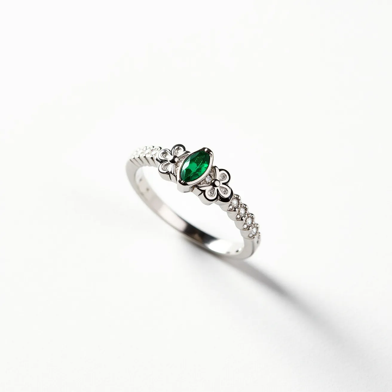 This emerald promise ring features a striking marquise-cut emerald set in a floral design. The central stone is flanked by two delicate flower motifs, enhancing its elegance and charm. The band is likely made of silver or white gold, adorned with small, round-cut diamonds set in a pave setting, which adds a subtle sparkle. The combination of the rich green emerald and the sparkling diamonds creates a captivating contrast, making this ring both elegant and meaningful.