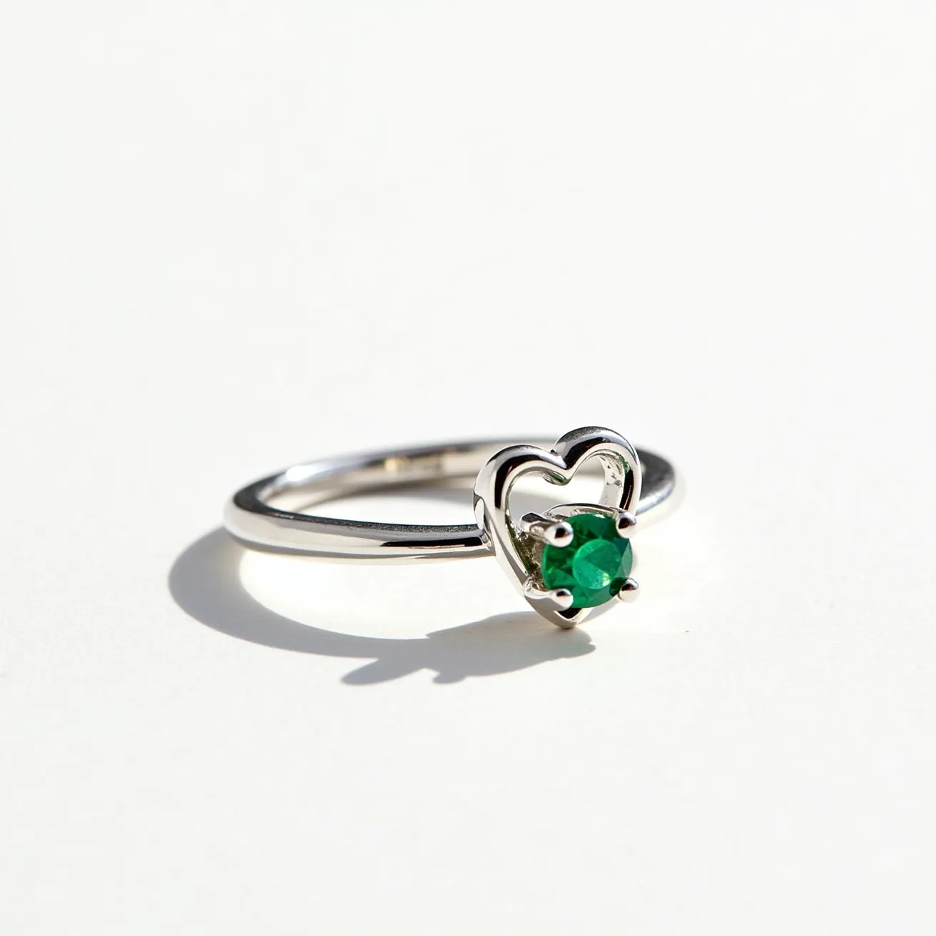 This emerald promise ring features a delicate silver band paired with a striking emerald gemstone. The emerald is round-cut, elegantly set in a prong setting nestled within a heart-shaped frame. The sleek metal band complements the vibrant green of the gemstone, creating a balanced and harmonious design. The heart-shaped setting adds a romantic element to the ring, while the prongs securely hold the emerald in place, ensuring both style and function.