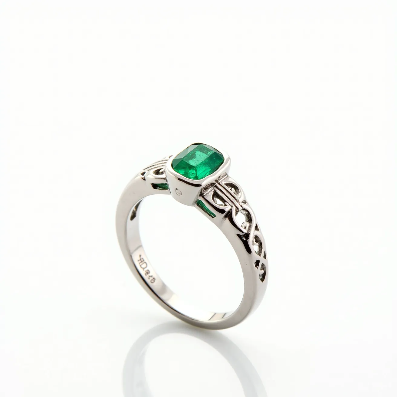 This emerald ring features a vibrant, cushion-cut emerald set in a sleek bezel setting. The band is made of a polished silver or white gold material, showcasing intricate Celtic-inspired detailing along the sides, adding an elegant and unique aesthetic. The smooth, reflective finish of the metal complements the rich green hue of the emerald, creating a striking contrast. There is no visible clasp, as the ring is a continuous band, designed for a secure and comfortable fit on the finger.