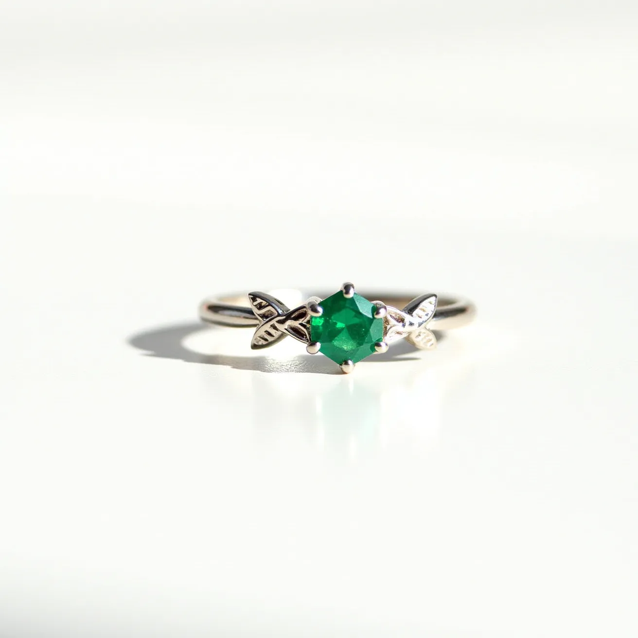 This emerald ring features a vibrant green emerald cut into a hexagonal shape, held securely by a six-prong setting that allows the gem to be the center of attention. The band is crafted from a polished metal, likely silver or white gold, which complements the deep green of the emerald. The ring is accented with intricate leaf motifs on either side of the stone, adding a touch of elegance and nature-inspired design to the piece. These details create a balanced and harmonious look, with the emerald drawing the eye and the subtle setting enhancing its beauty.