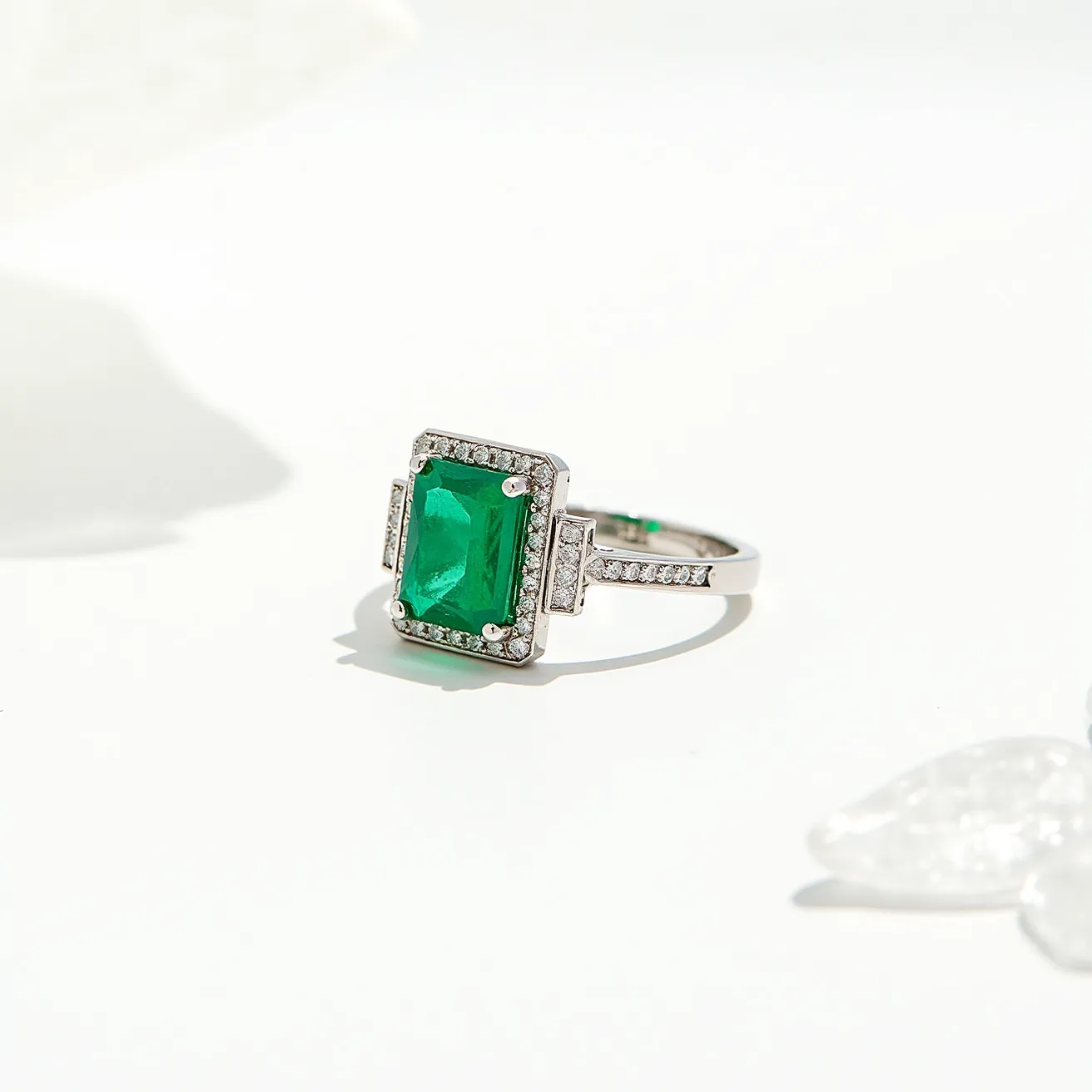 This emerald ring features a stunning rectangular-cut emerald at its center, set in an elegant four-prong setting. The central stone is surrounded by a halo of small, round clear gemstones, likely diamonds, that enhance the vibrant green of the emerald. The band appears to be made of polished white metal, potentially platinum or white gold, which complements the brightness of the stones. The shoulders of the ring are also adorned with tiny diamonds, adding to its luxurious and intricate design. The craftsmanship displays attention to detail, with the prongs and setting securely cradling the emerald, ensuring both beauty and durability.