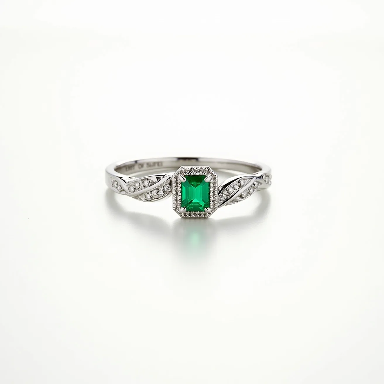 This emerald ring features a vibrant, rectangular-cut emerald set within a sophisticated white metal band, likely platinum or white gold. The stone is securely held in a bezel setting, which is surrounded by a delicate halo of small, sparkling diamonds, enhancing its rich green hue. The band showcases intricate detailing with a series of small diamonds on each side, artfully arranged in a twisting design. There is no visible clasp or attachment, as it is a classic ring design meant to slide onto the finger and be worn securely, combining elegance with modern sophistication.