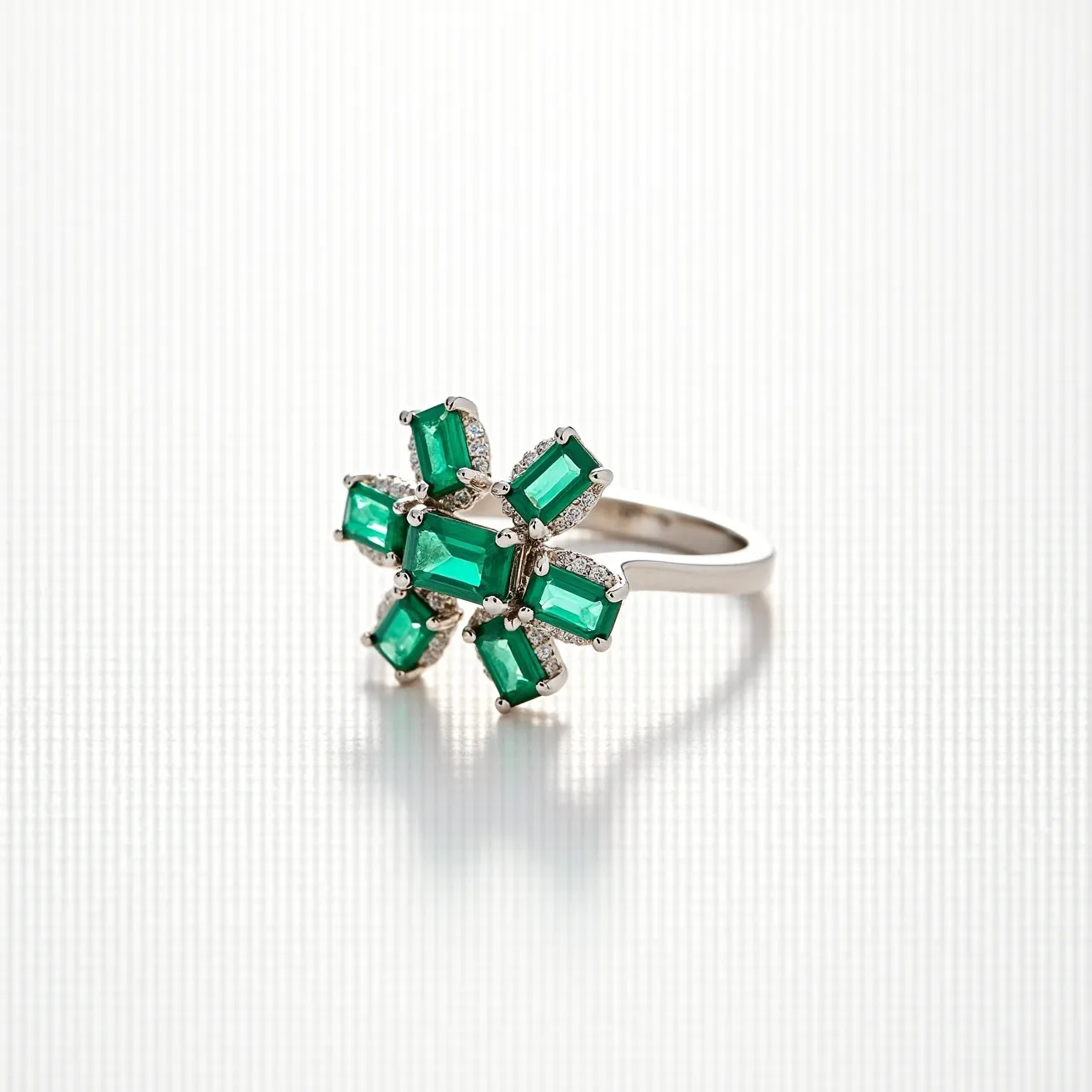 This emerald ring features a striking floral design, composed of multiple rectangular-cut emeralds arranged symmetrically around a central point. Each stone is securely set in a prong setting that enhances their vibrant green hues. Surrounding the emeralds are small, brilliant-cut diamonds, adding a touch of sparkle and contrast. The band of the ring, likely made of white gold or platinum, complements the overall design with its smooth, polished finish. The craftsmanship highlights the beauty and elegance of the gemstones, making it a standout piece of jewelry.