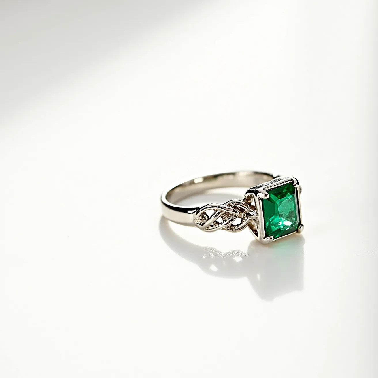 This emerald ring features a prominent rectangular emerald stone set in a four-prong setting, which elegantly showcases its vibrant green hue. The band is crafted from a lustrous metal, likely white gold or platinum, adding a sleek and sophisticated finish to the piece. Enhancing the band is an intricate knot design, adding a touch of elegance and complexity to the overall aesthetic. The combination of the rich emerald stone with the detailed band offers a classic yet sophisticated look, perfect for a range of occasions.