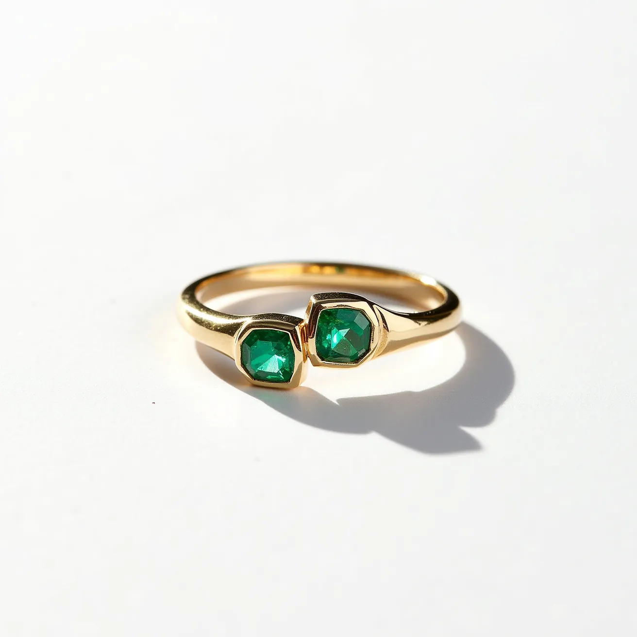 This emerald ring features two vivid green emerald gemstones, each cut into a hexagonal shape. The stones are set in a sleek bezel setting, which provides a modern and secure fit. The band appears to be made of polished yellow gold, complementing the rich green hue of the emeralds and adding a classic touch to the overall design. The ring's minimalist and elegant structure highlights the beauty of the materials and craftsmanship.