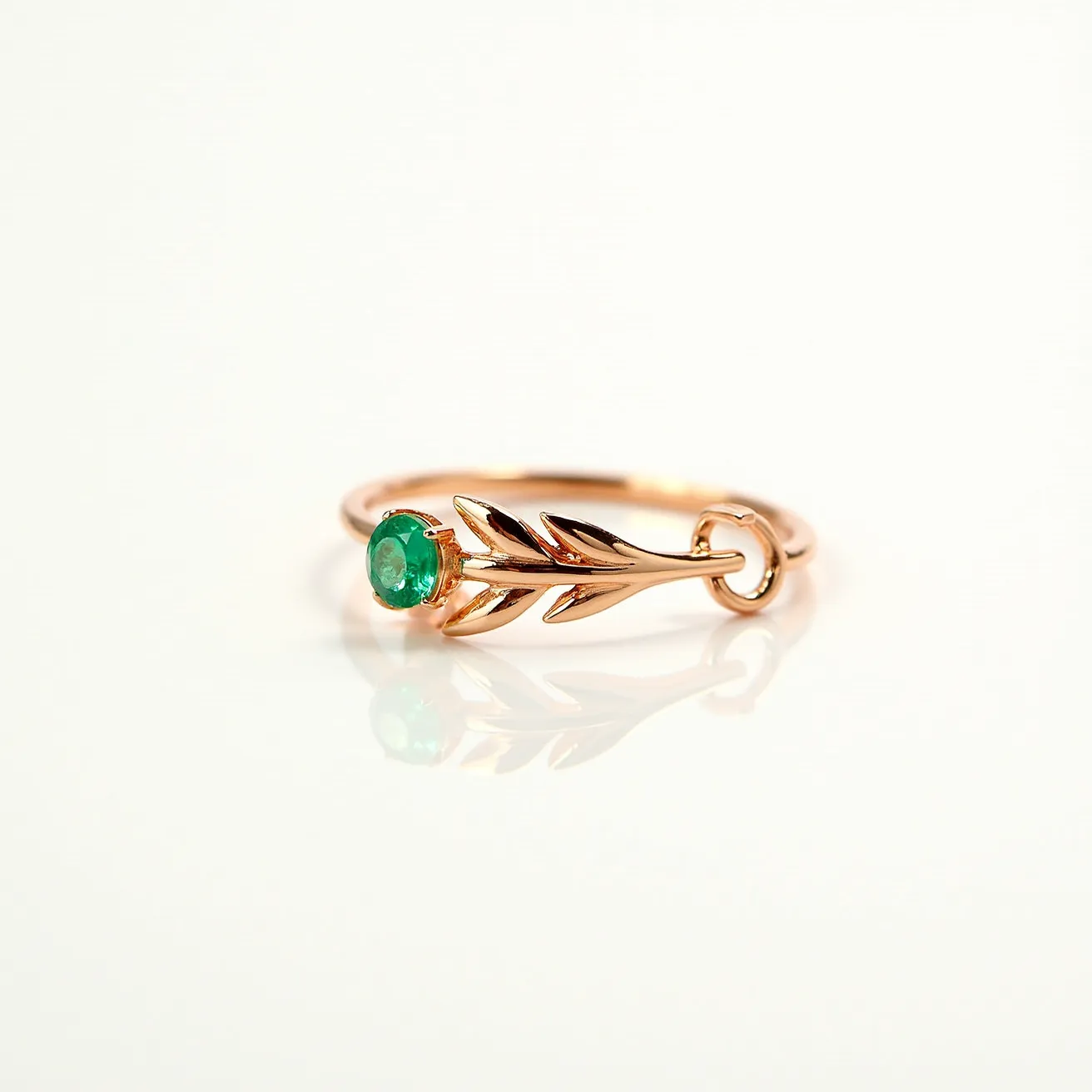 This emerald ring features a central round-cut emerald stone set in a prong setting, highlighting its vibrant green hue. The band is crafted from a gold-toned metal, designed with elegant, leafy details that curve gracefully around the gemstone. The intricate leaf motif adds an artistic element to the overall design, enhancing its organic and nature-inspired appearance. The ring lacks any visible clasps or additional attachments, presenting a seamless and delicate aesthetic.